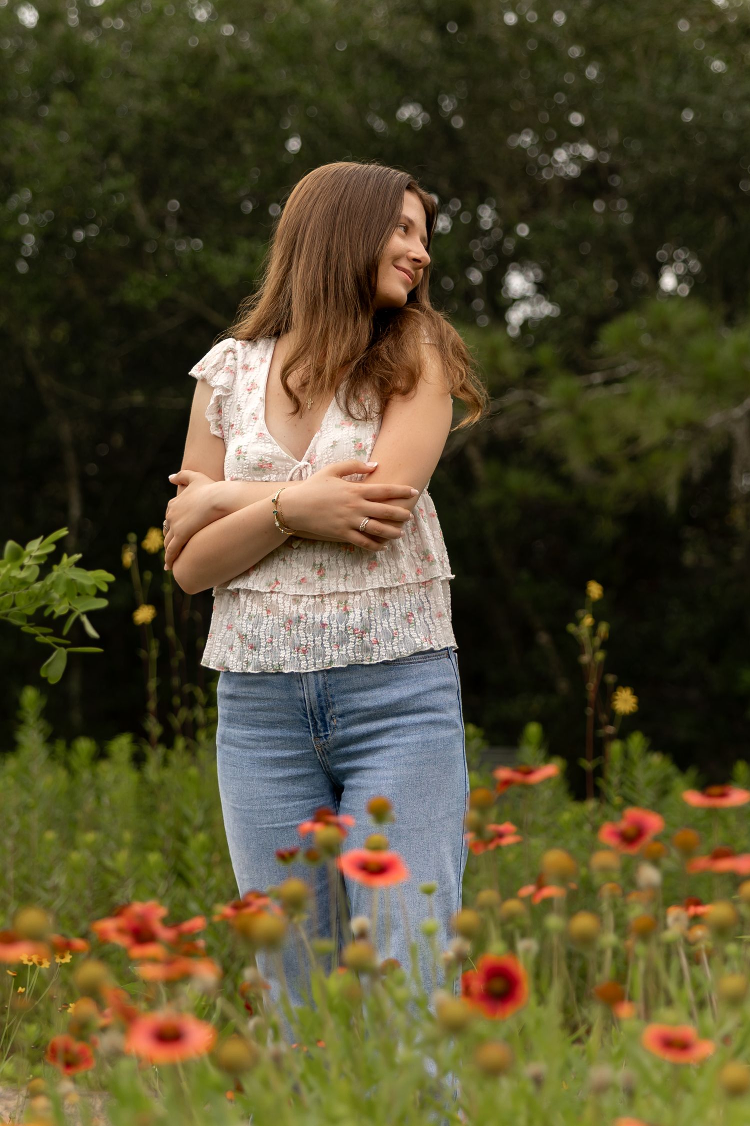 Young woman in floral top and jeans standing in a field of red wildflowers, smiling and looking away.
