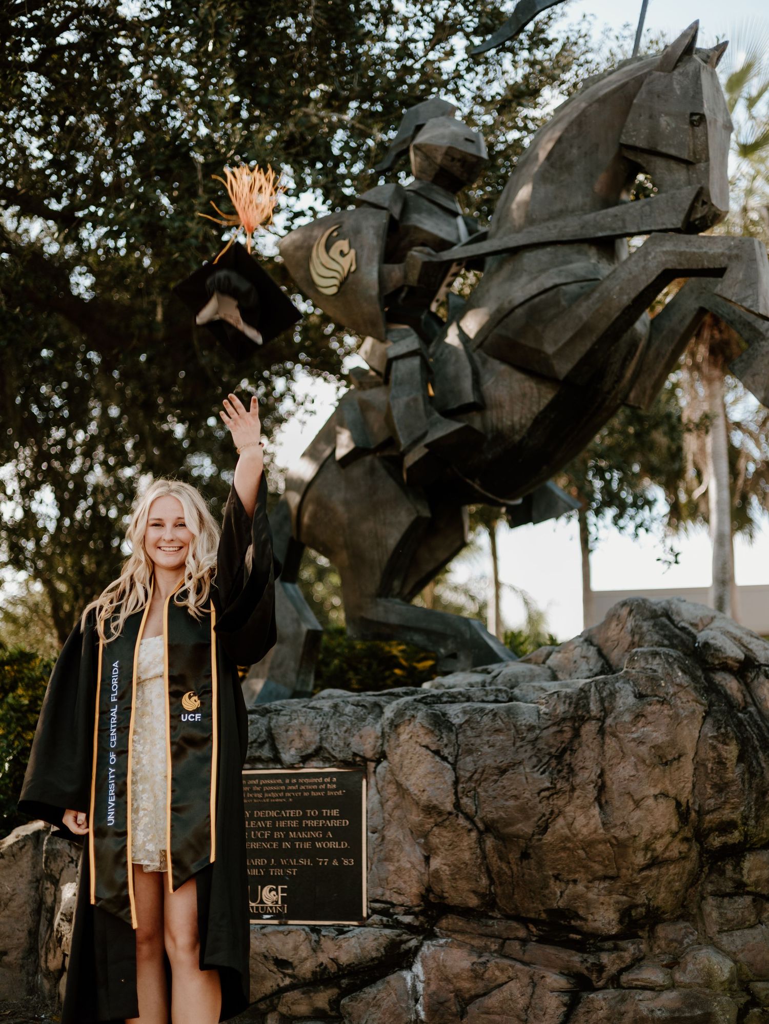 A smiling female graduate in black and gold regalia poses by a knight on horseback statue at UCF.