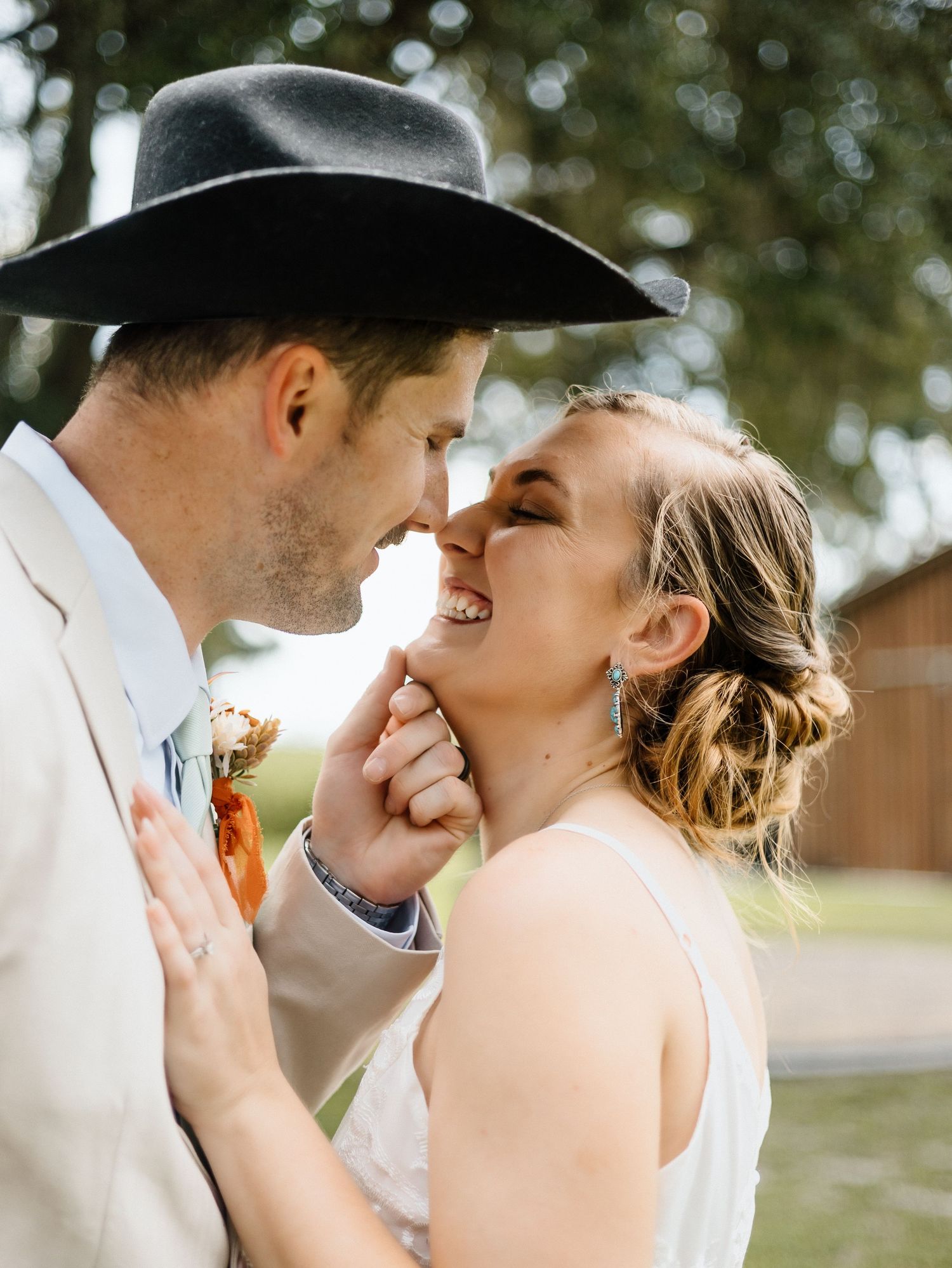 A joyful bride and groom share a romantic moment, the groom wearing a black cowboy hat and boutonniere at their wedding.