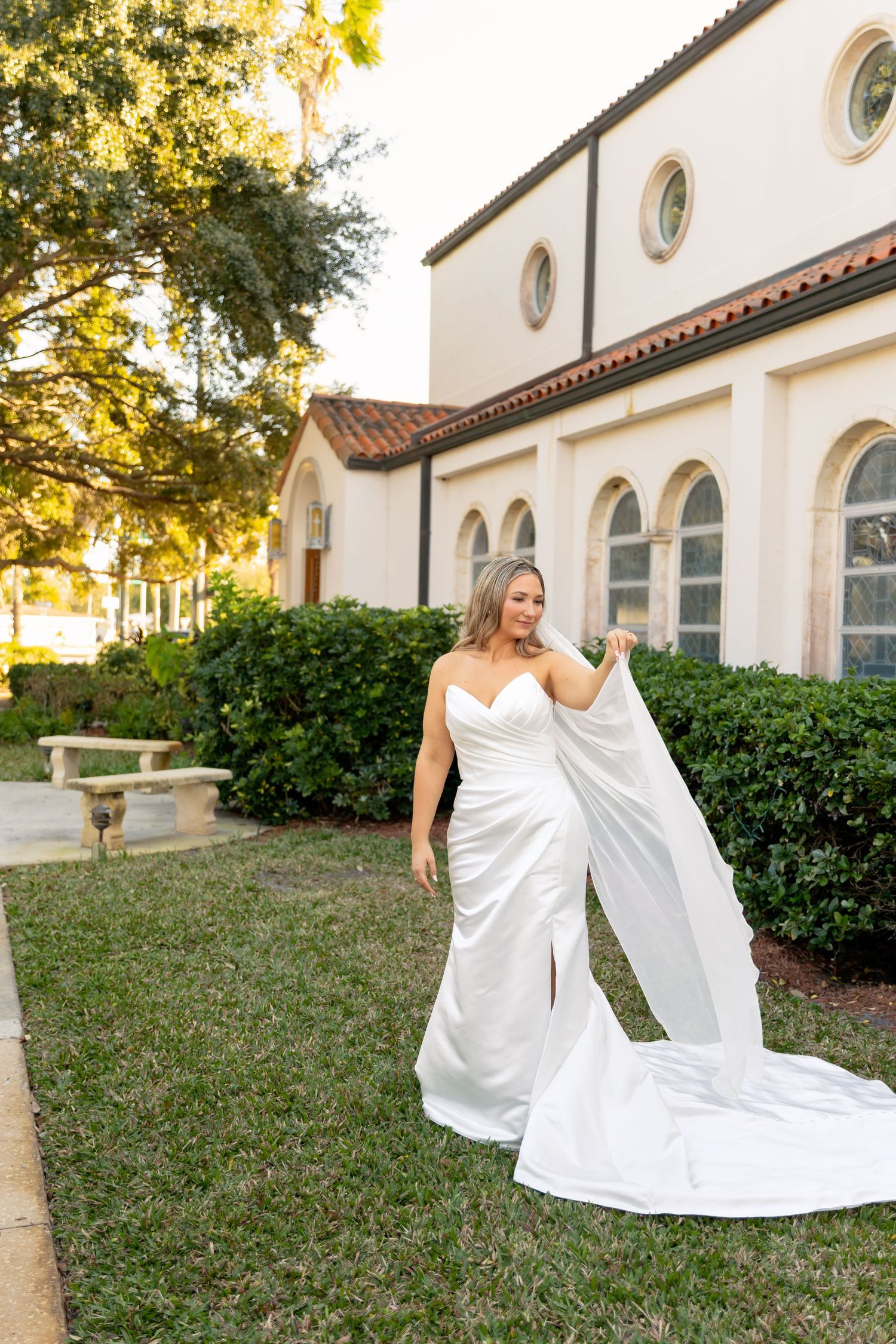 Bride in white strapless gown holding flowing train outside elegant Spanish-style building with arched windows.