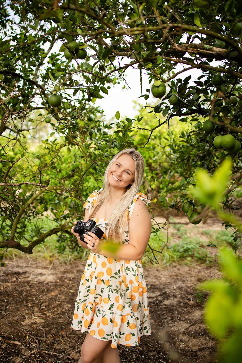 Smiling blonde woman in orange-print sundress holding a camera while standing among lush green citrus trees.