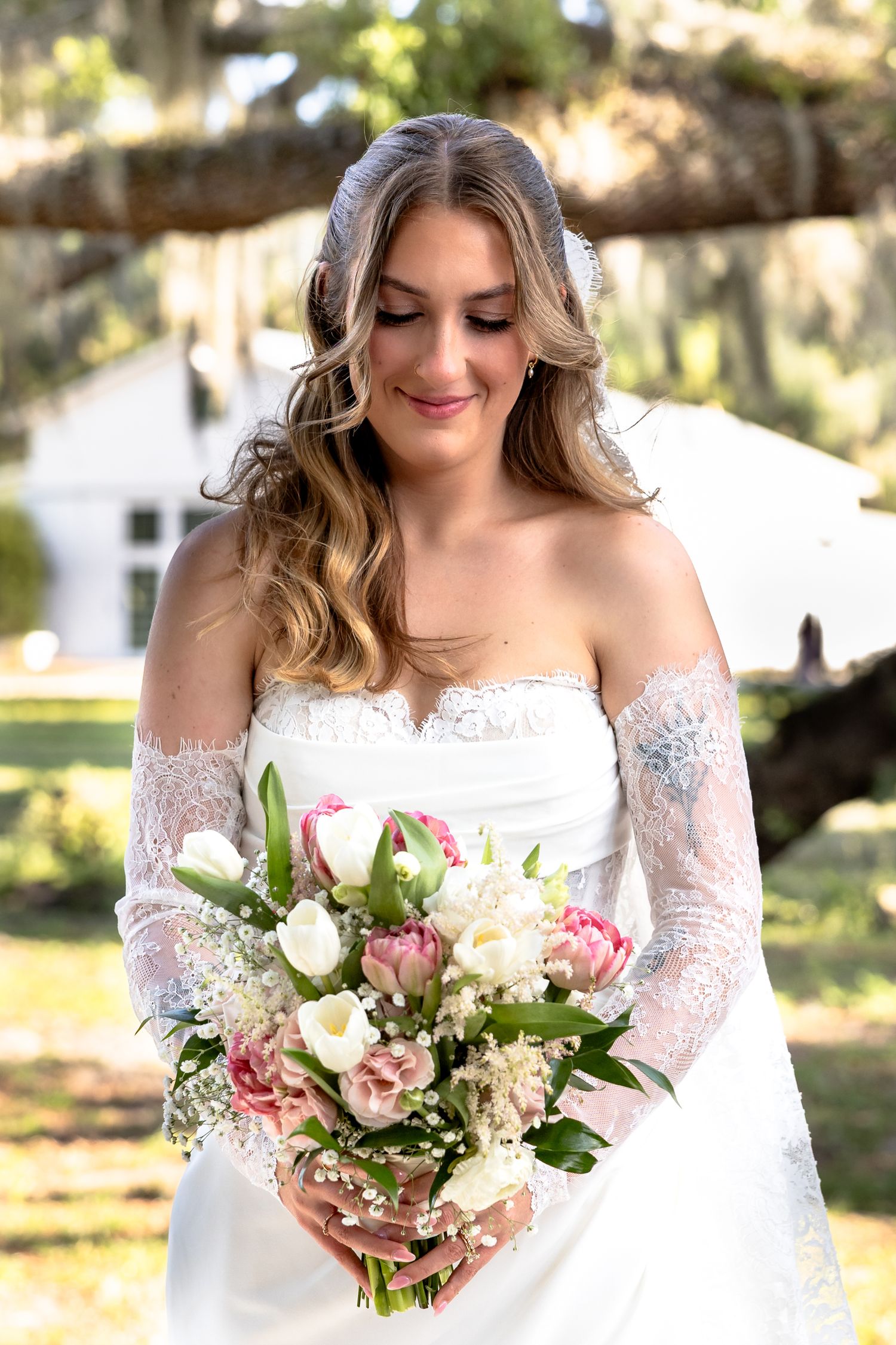 Bride in lace off-shoulder gown holding pink and white tulip bouquet, smiling outdoors under moss-draped trees.