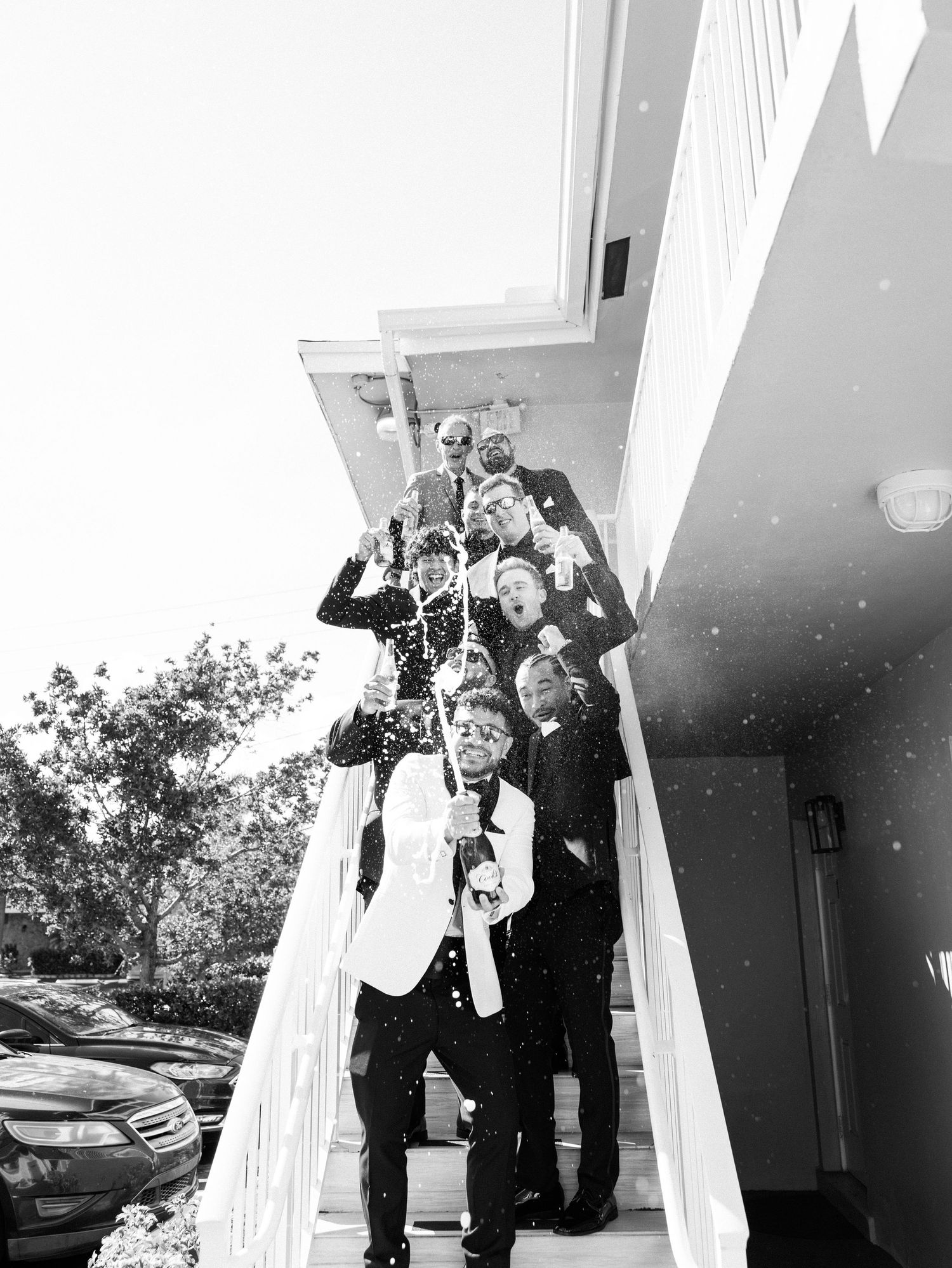 Groomsmen in tuxedos celebrating on stairs with champagne spray in black and white wedding photo.