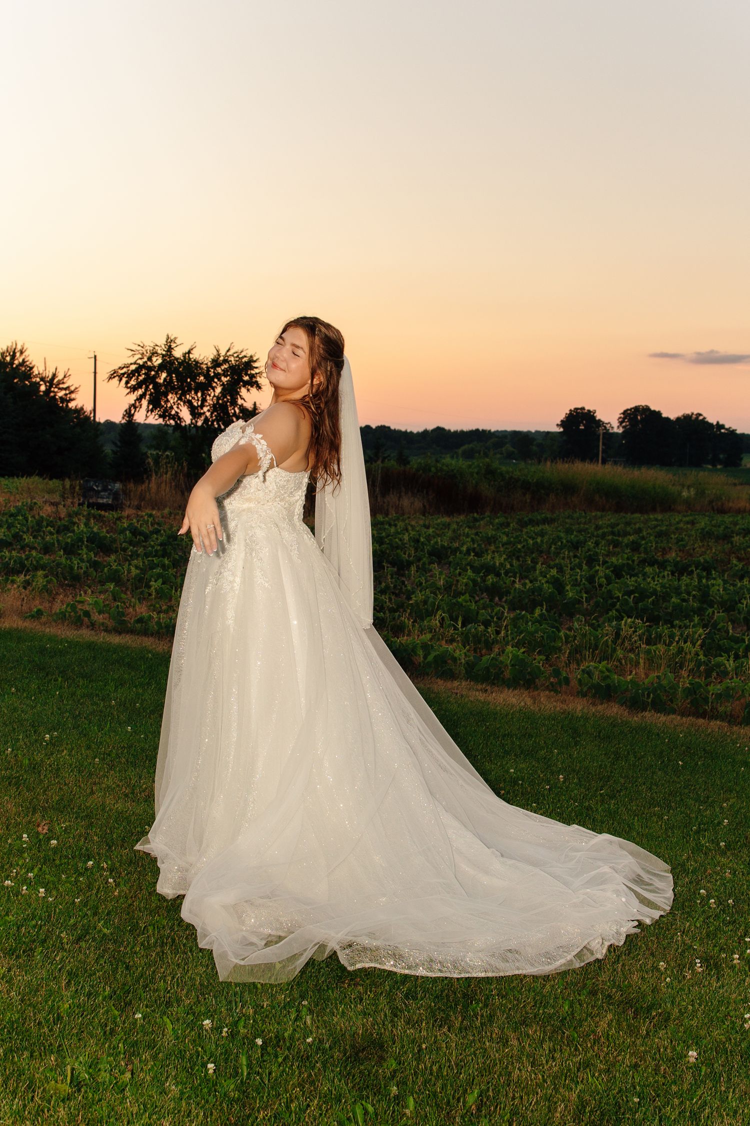 Bride in white tulle gown with veil poses gracefully outdoors at sunset near vineyard.