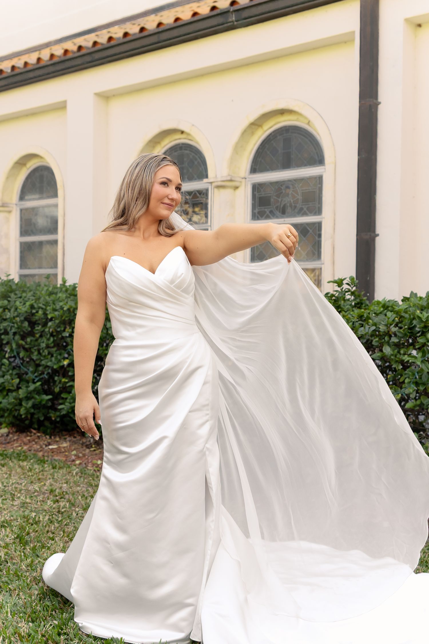 Bride in strapless white satin wedding gown lifting her flowing train outdoors near a yellow stucco building.