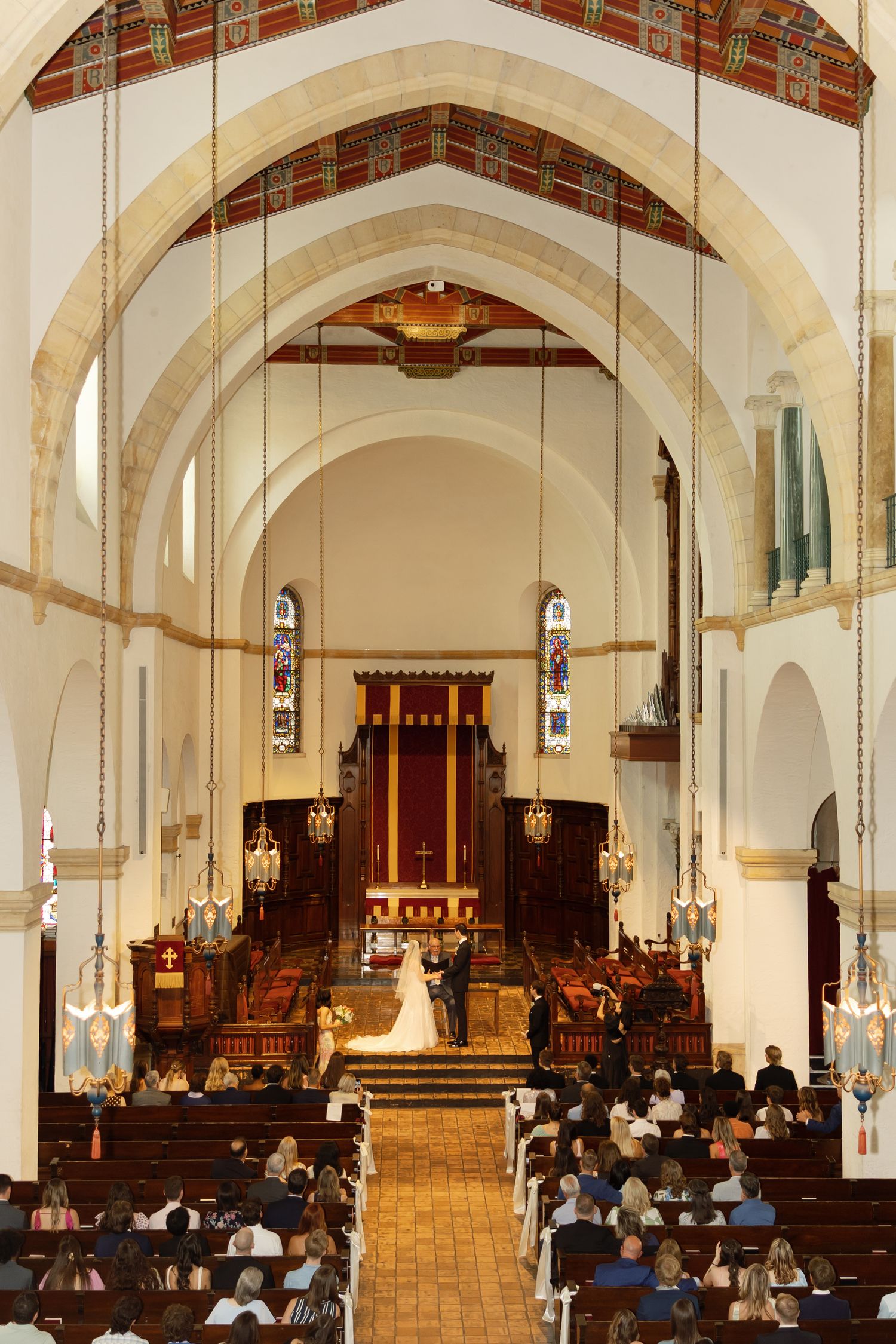 Wedding ceremony inside a grand church with arched ceilings, stained glass windows, and guests seated in wooden pews.