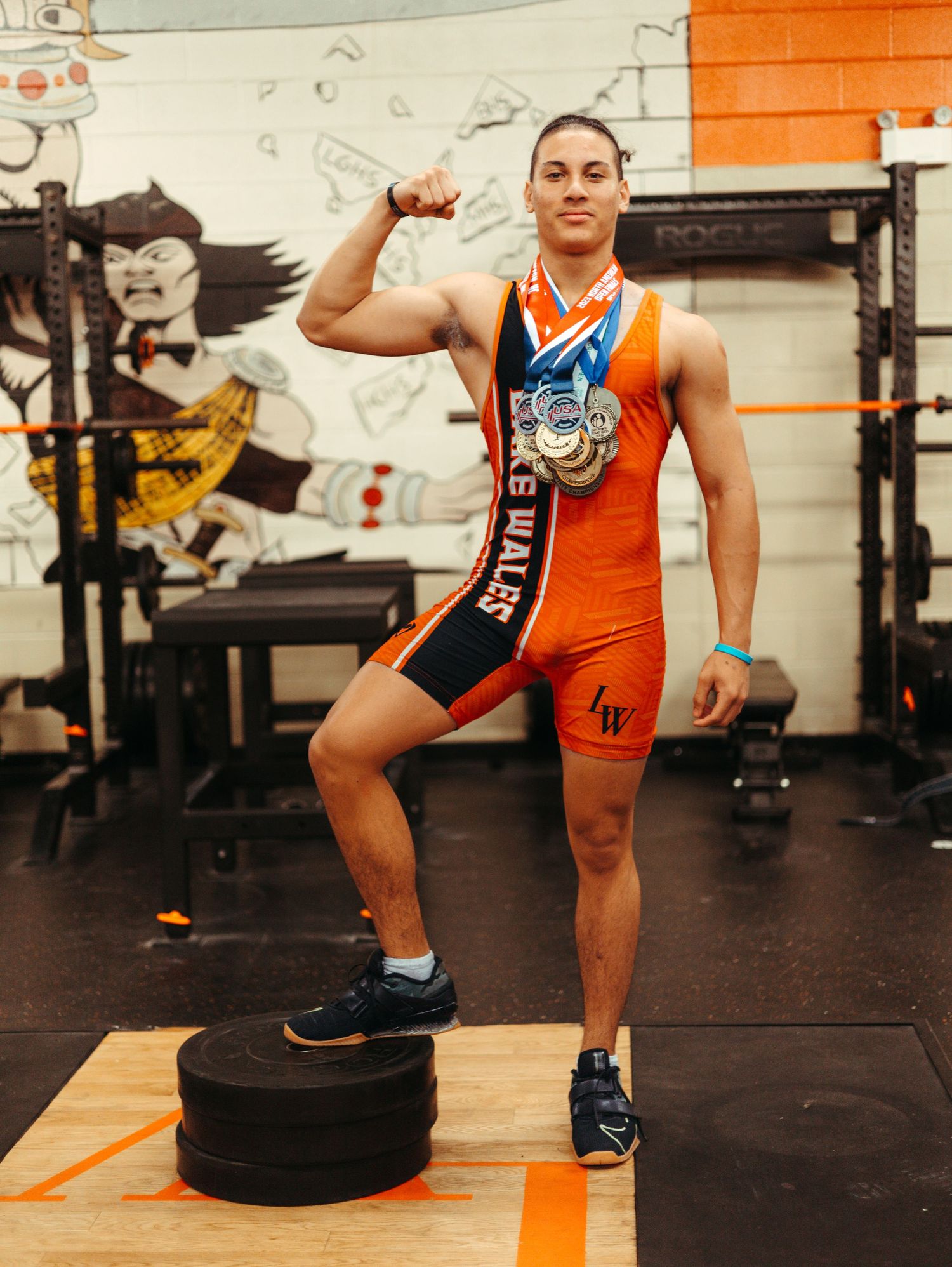 Athlete in orange singlet strikes victory pose on weight plates showing medal.