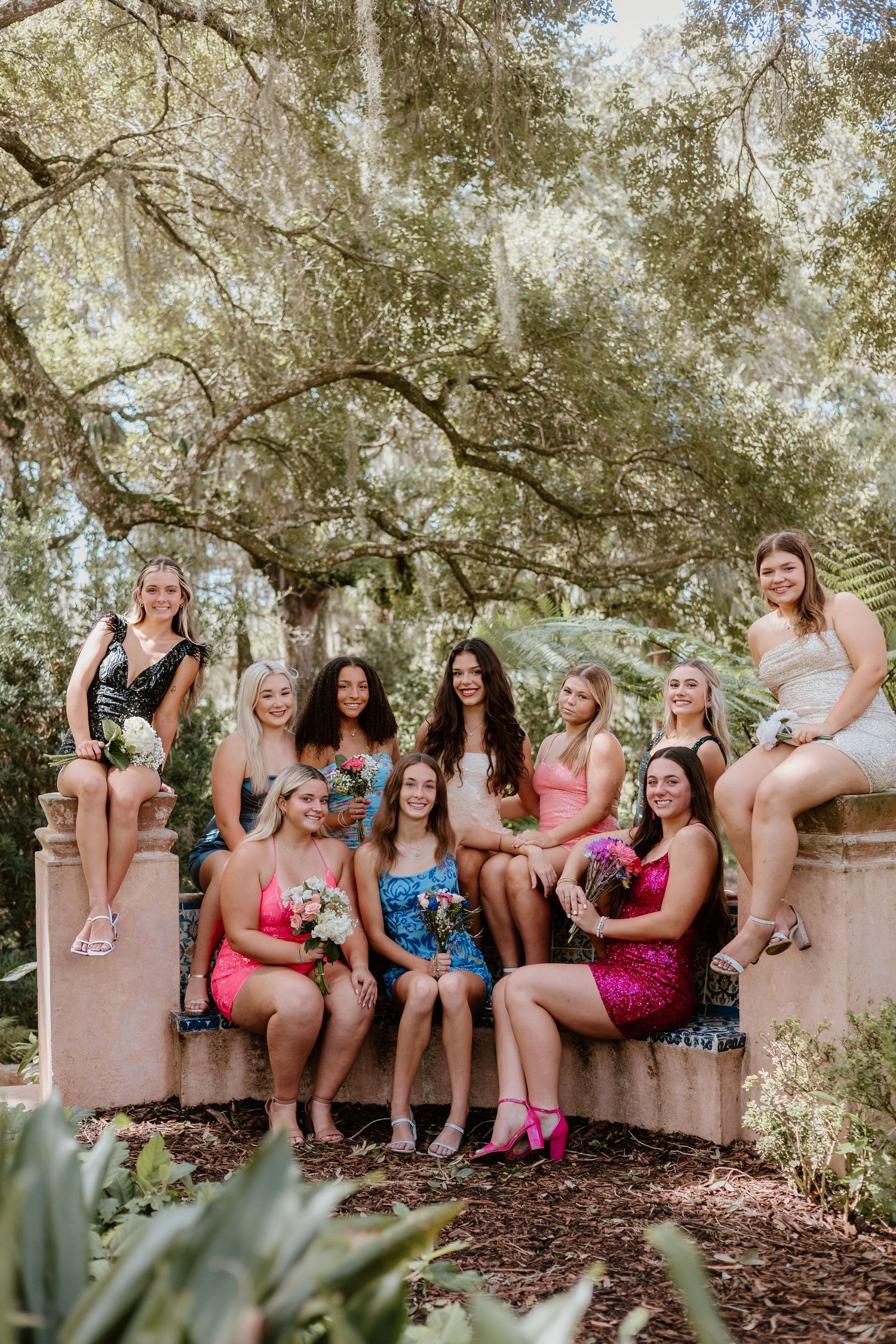 Wedding party in pink dresses sits on stone wall under shady trees capturing candid laughter-filled moments.