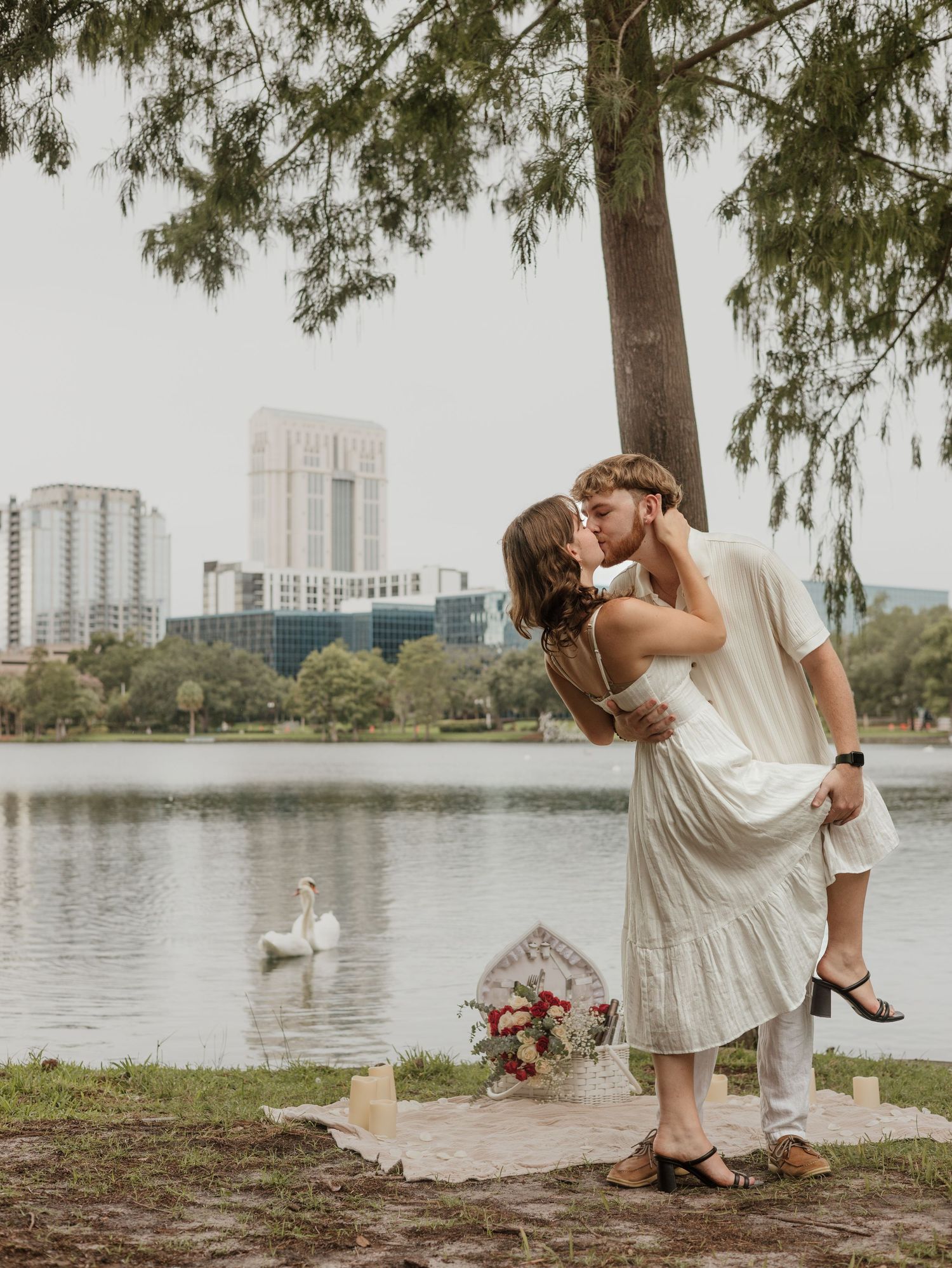 Romantic couple shares an intimate moment by a lake in a park setting with city buildings in the background and swans swimming.