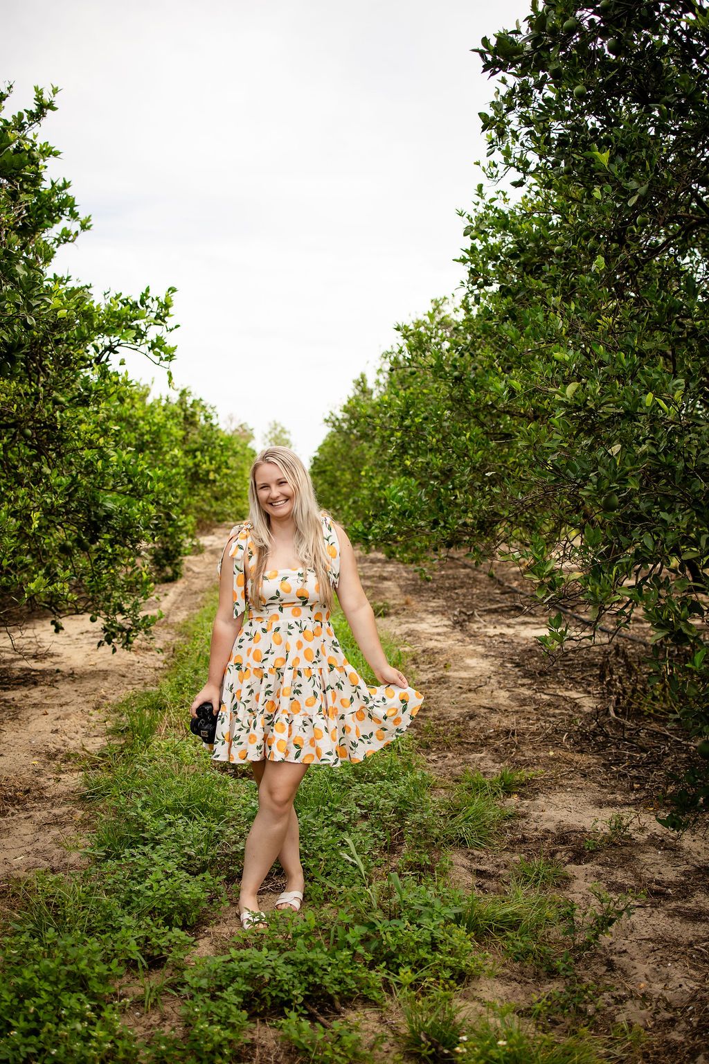 Smiling blonde woman in floral dress standing in lush green orchard pathway on overcast day.