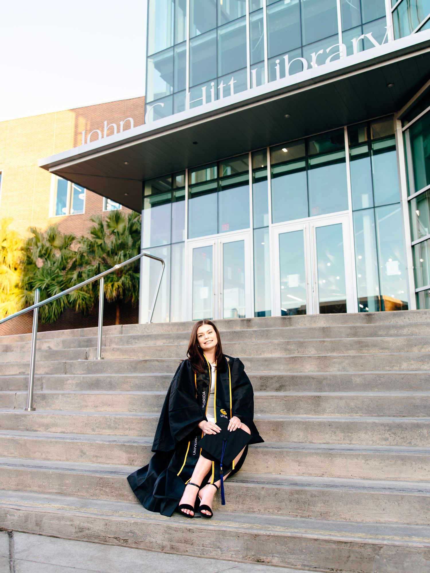Graduate in black cap and gown sits on steps outside John Lil Library, looking up thoughtfully.