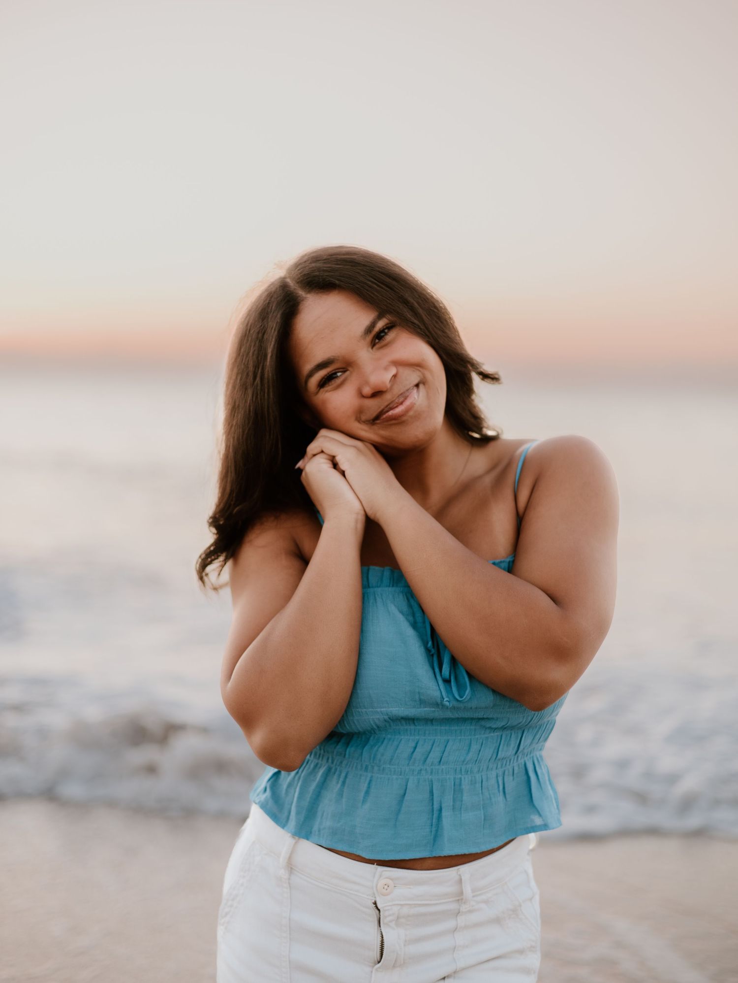 A person in a turquoise top and white pants poses joyfully on a beach at sunset.