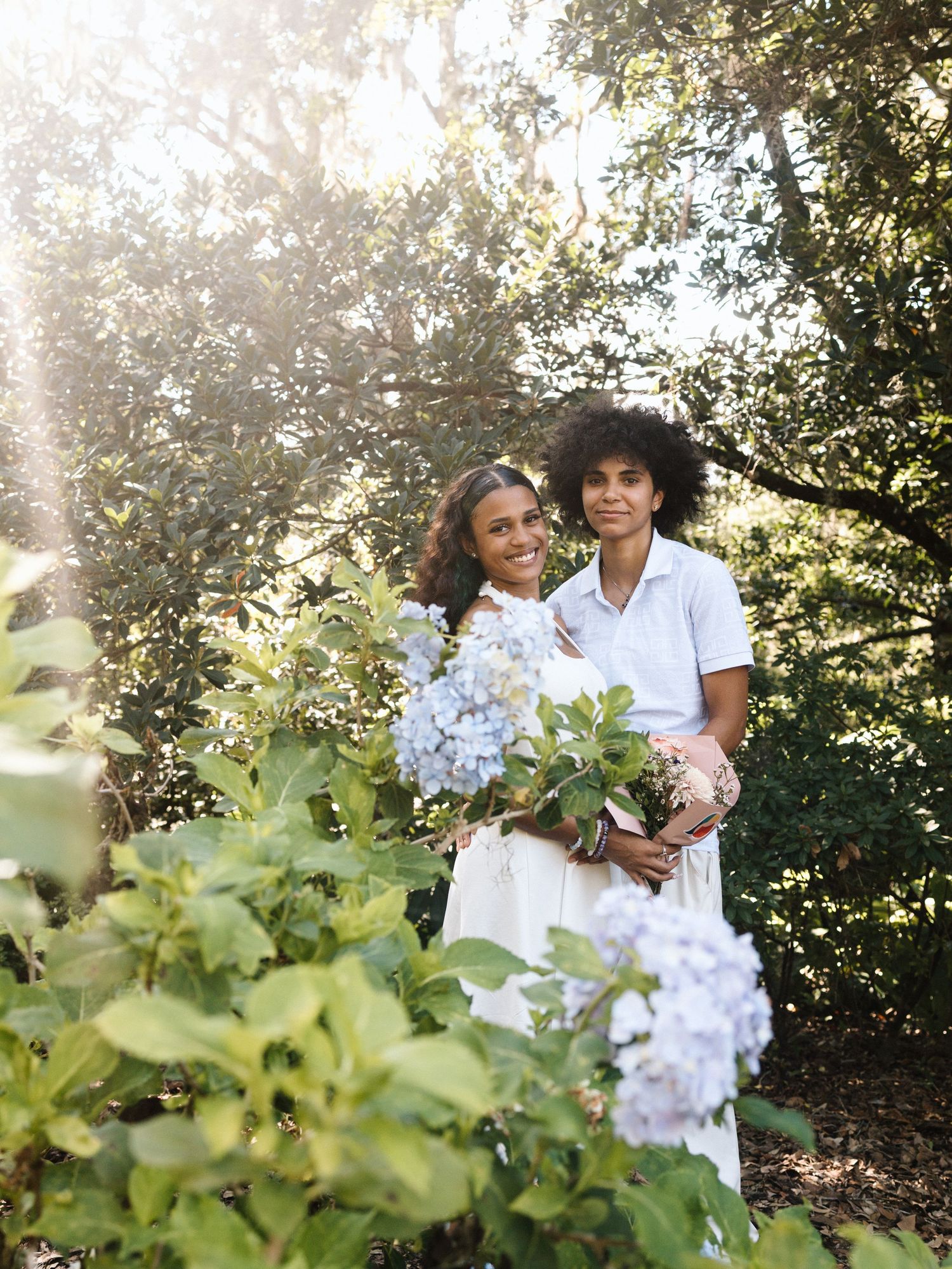 A couple holds blue hydrangeas while posing in a sunlit garden.