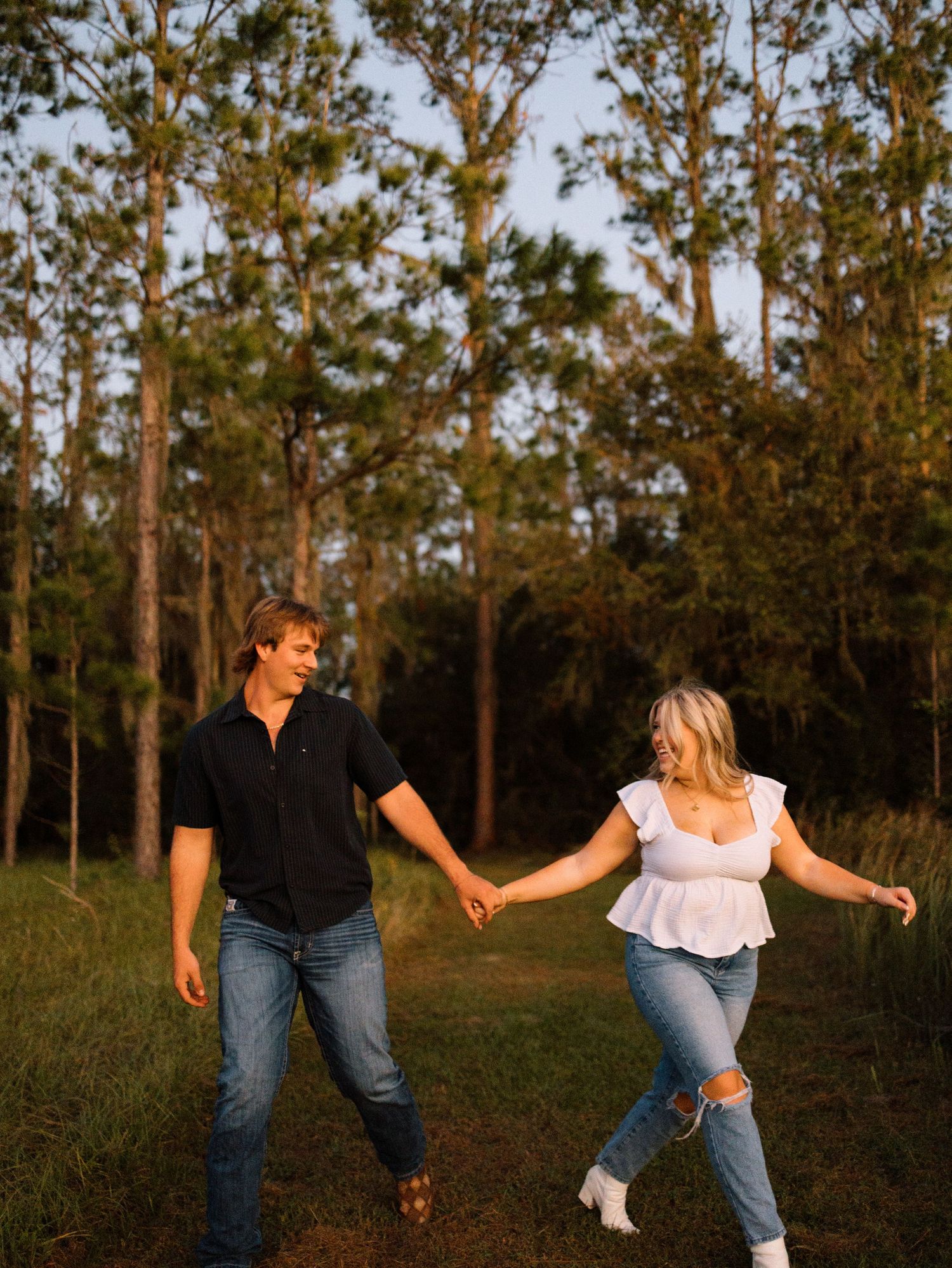 Dancing couple in denim and casual wear twirling and having fun in a natural pine forest setting at sunset.