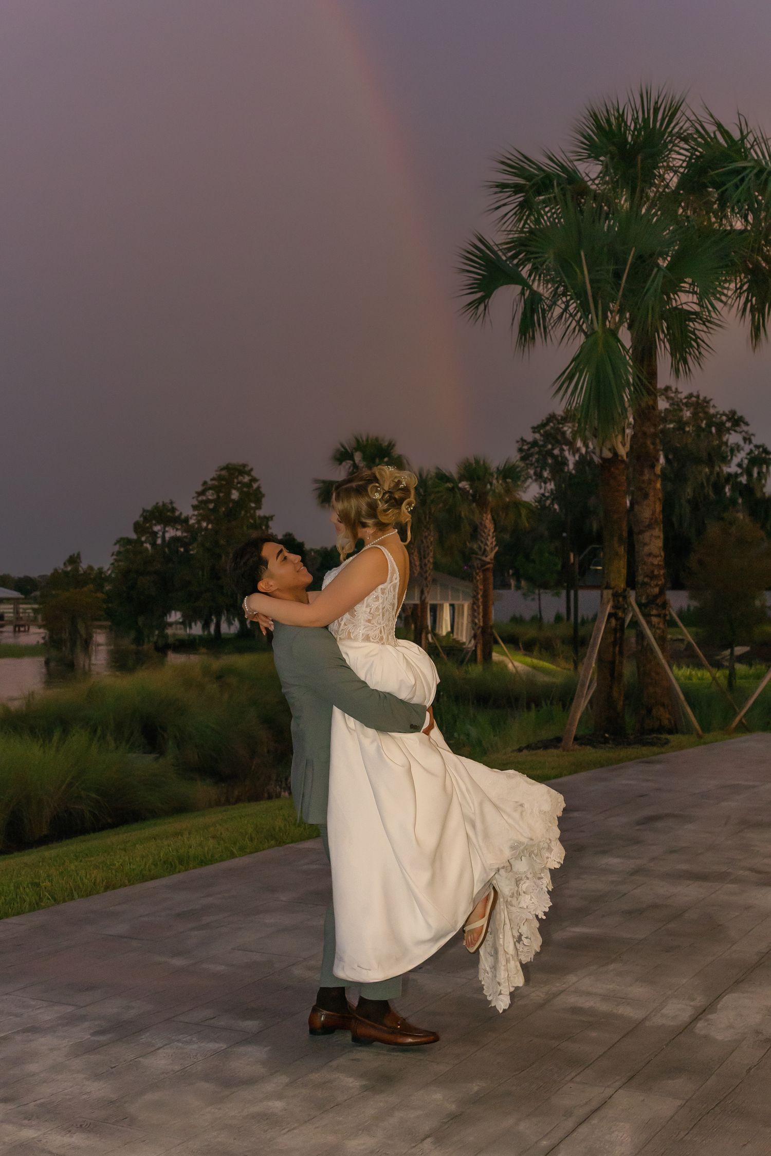 Groom lifting bride in white dress outdoors with a rainbow over palm trees at dusk wedding celebration.