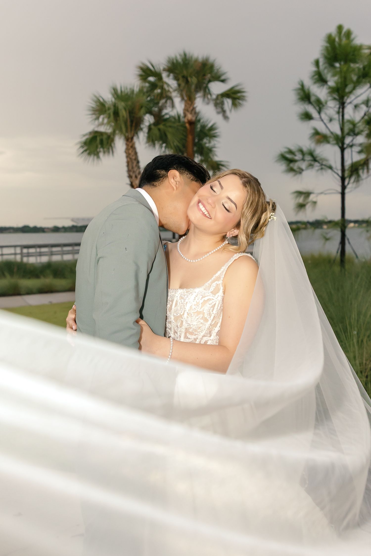 A bride in a lace gown smiles as her groom kisses her cheek, with a flowing veil and palm trees in the background.