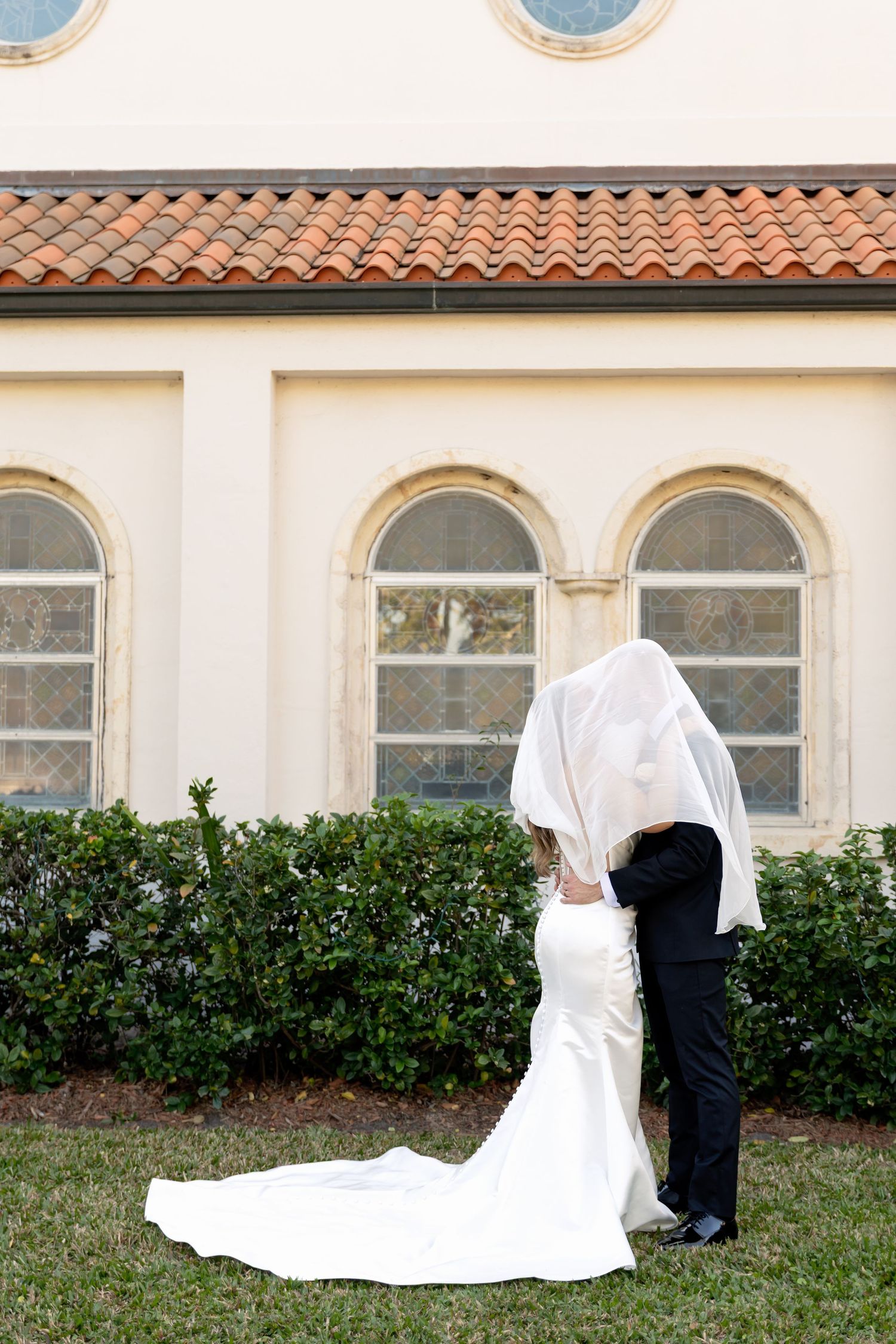 Bride and groom sharing a kiss under a white veil outside an elegant building with arched windows and terracotta roof.