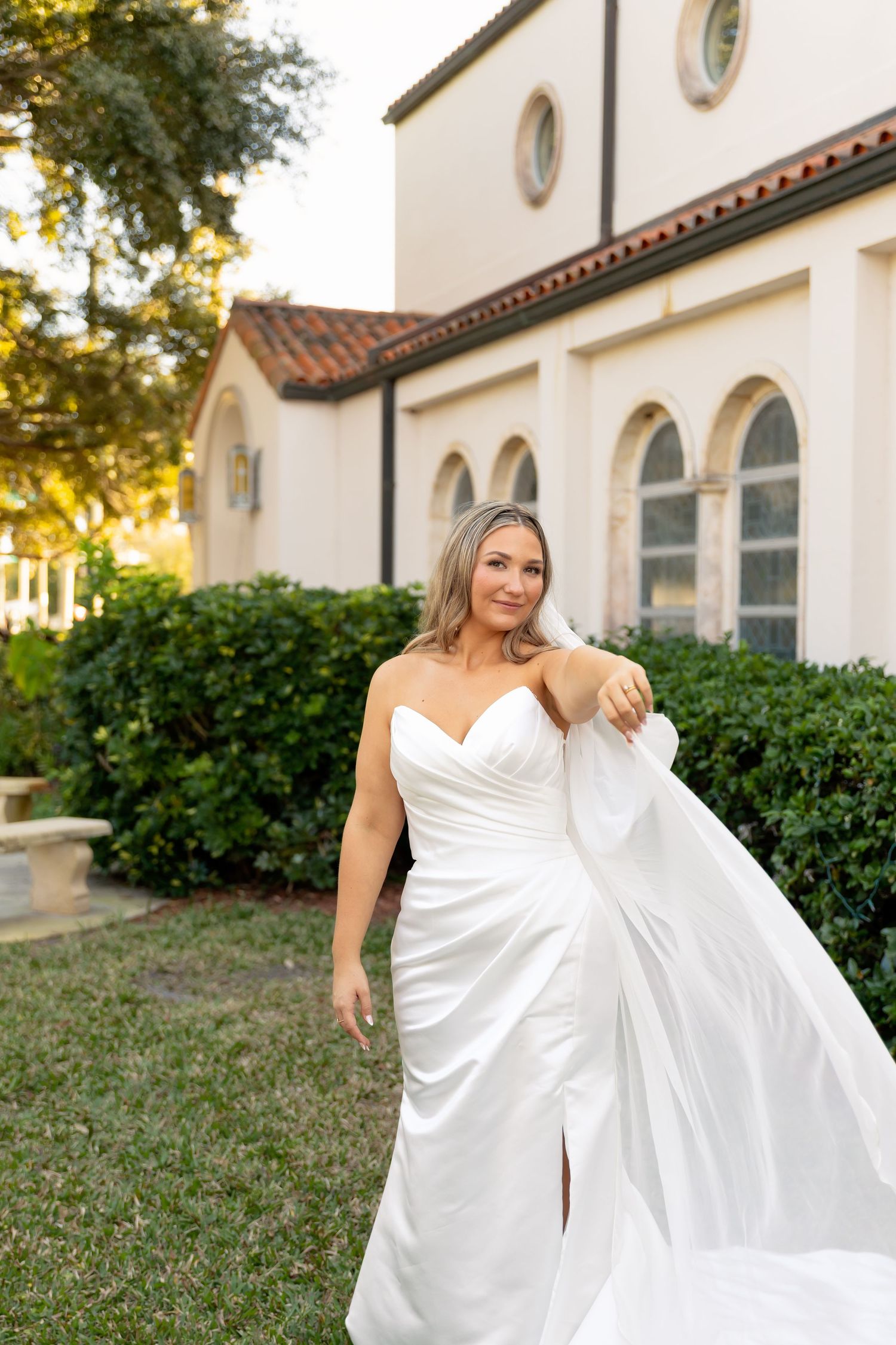Bride in strapless white satin gown holding veil outside elegant Spanish-style building with arched windows.