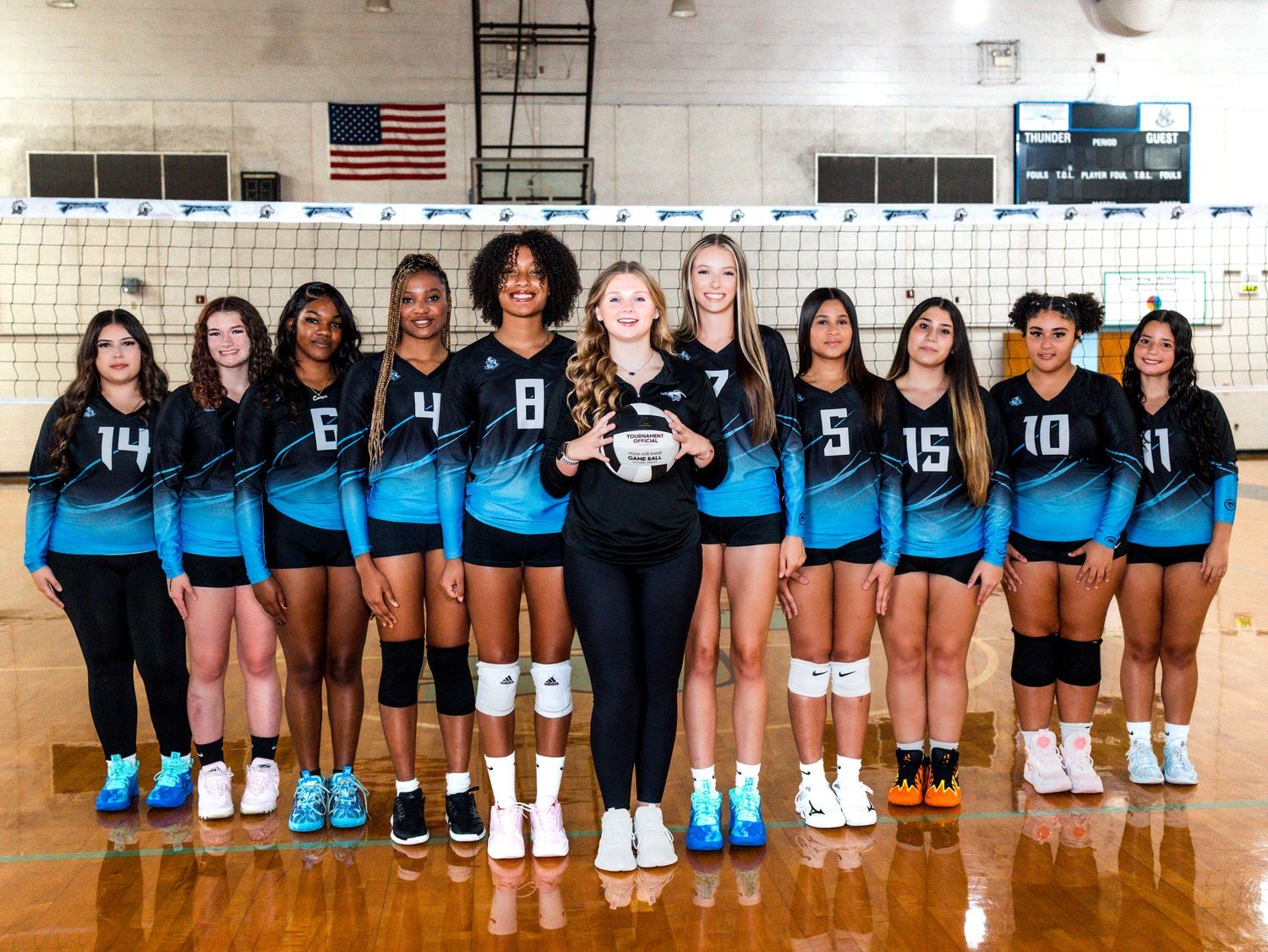 High school volleyball team in bright blue jerseys poses together on court with American flag in background.