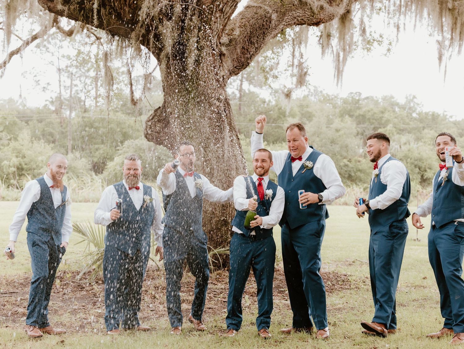 Groomsmen spraying champagne in celebration under a large oak tree.