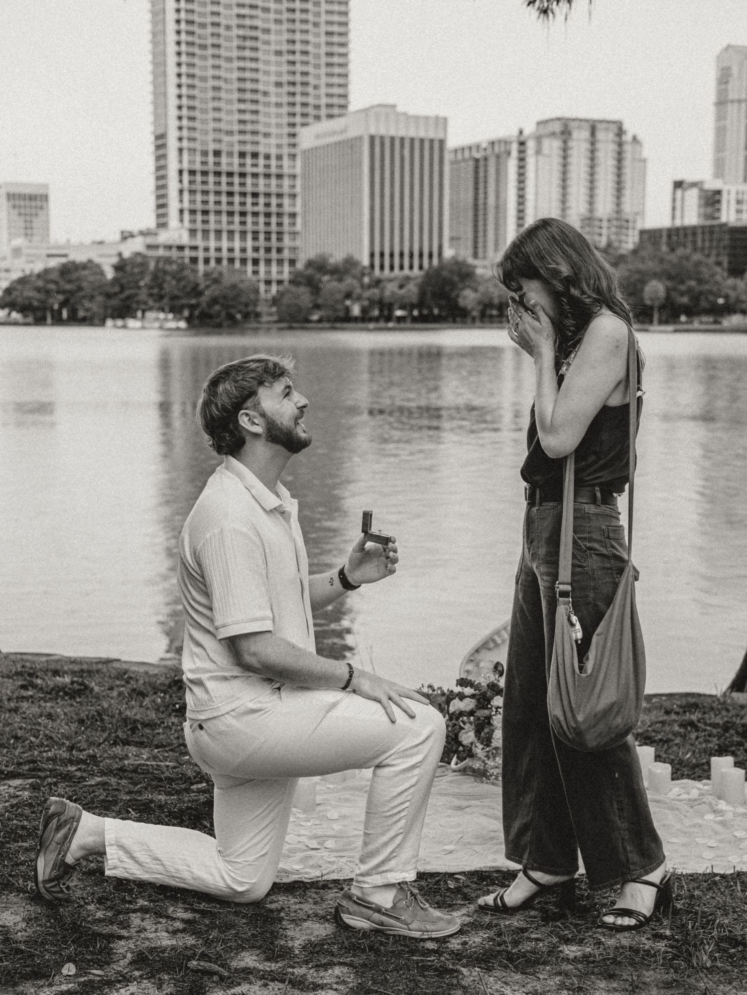 A black and white photograph shows a marriage proposal taking place by a lake with a city skyline in the background.
