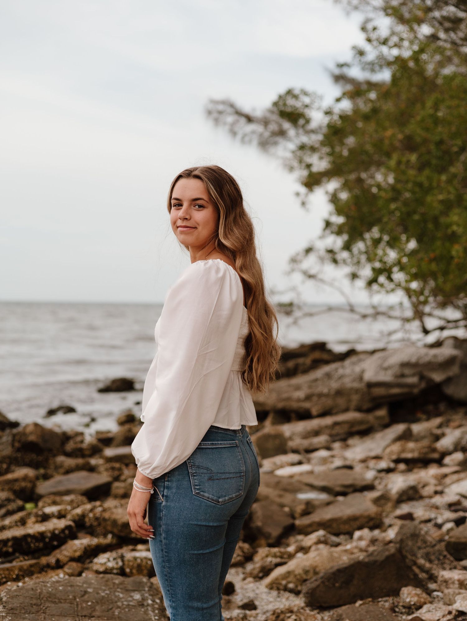 A person in a white blouse and jeans stands on rocky coastal terrain with ocean and trees in the background.