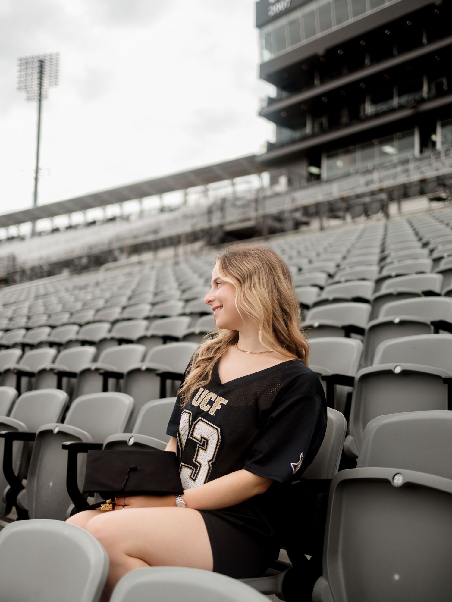 A smiling young woman in a black UCF jersey sits in empty stadium bleachers, holding a graduation cap.