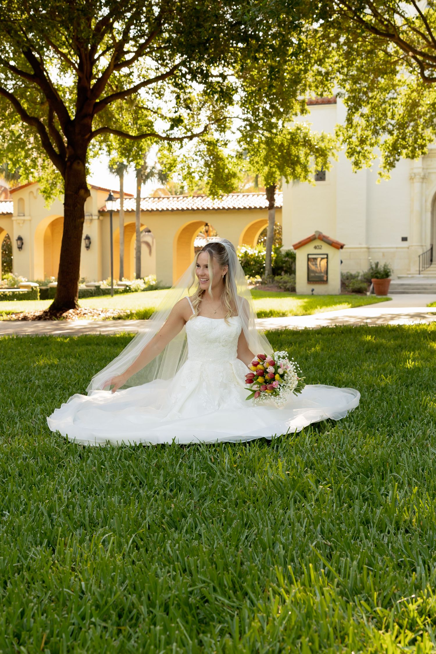 Bride in white gown sitting on lush green grass holding colorful bouquet, with elegant building and trees in background.