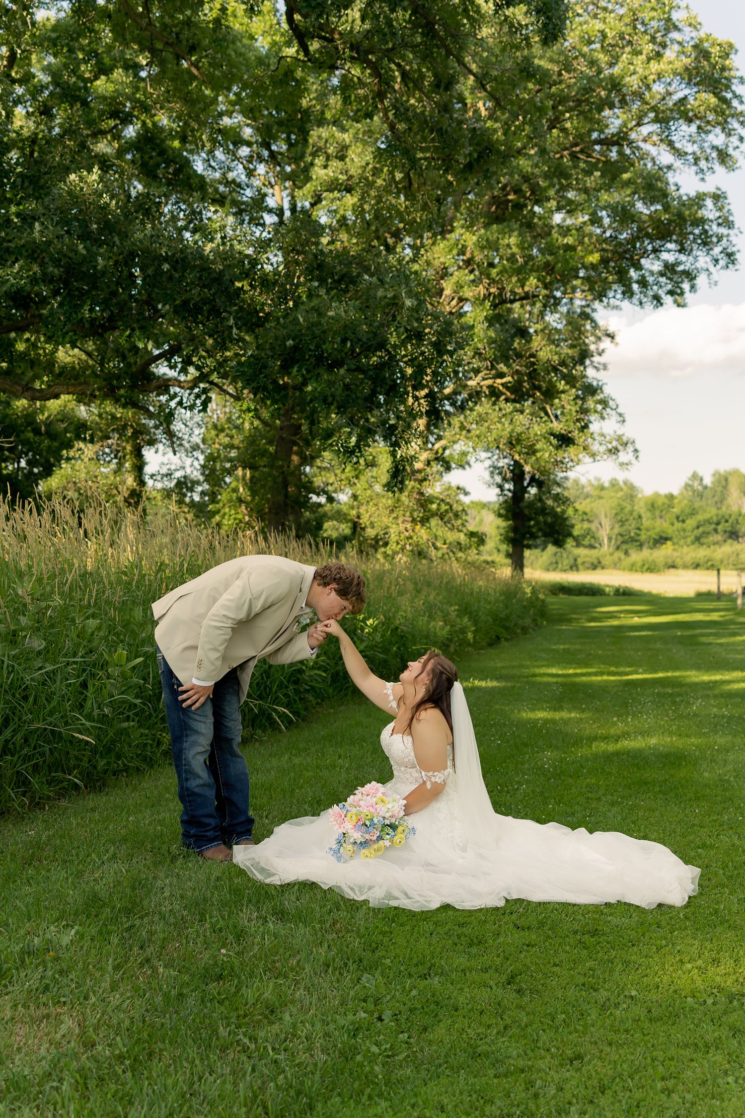 Bride in white gown sits on grass while groom in tan jacket leans down for a kiss outdoors.