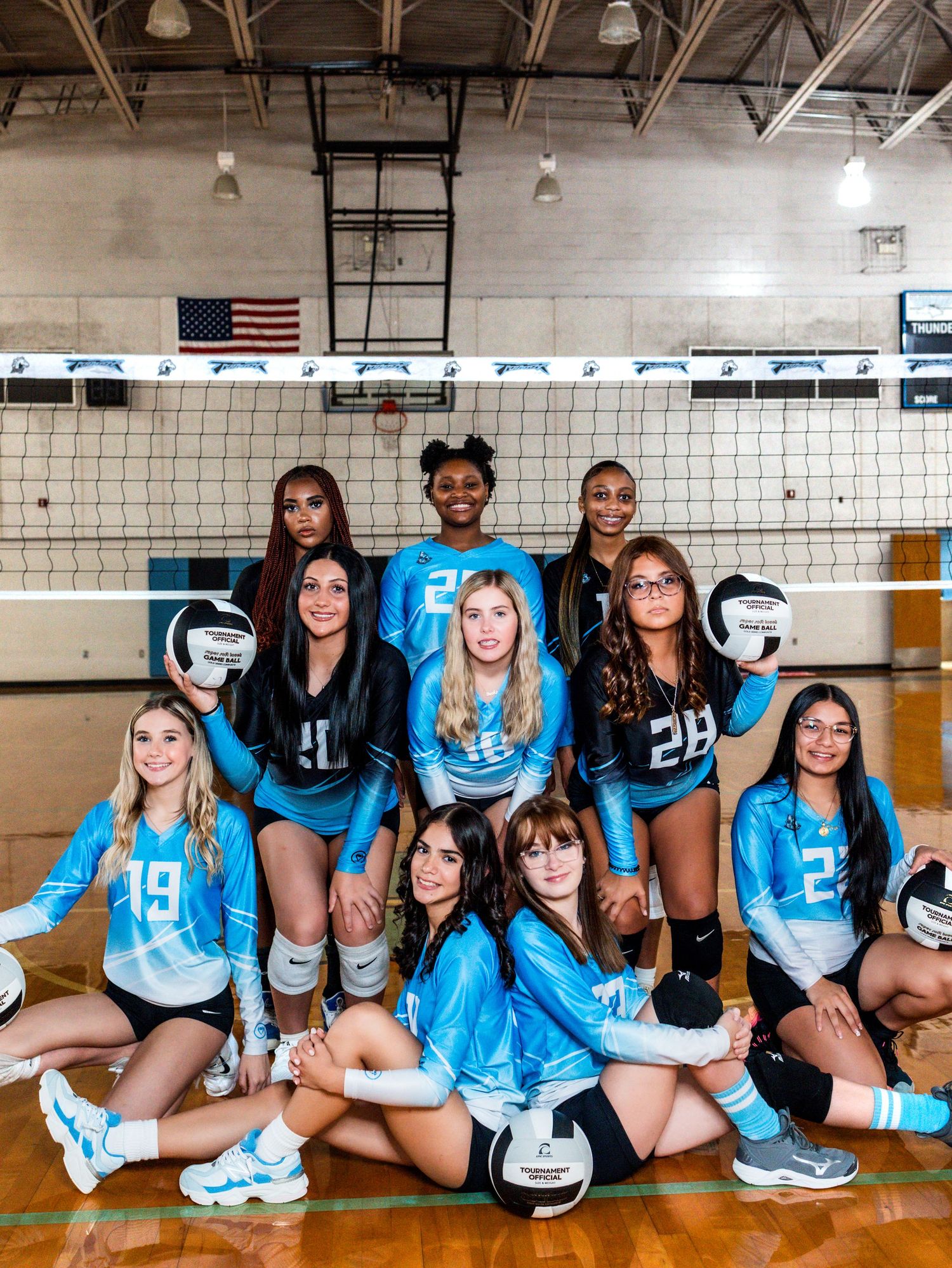 Members of a volleyball team wearing bright blue uniforms sit together on a gymnasium floor for a team photo.