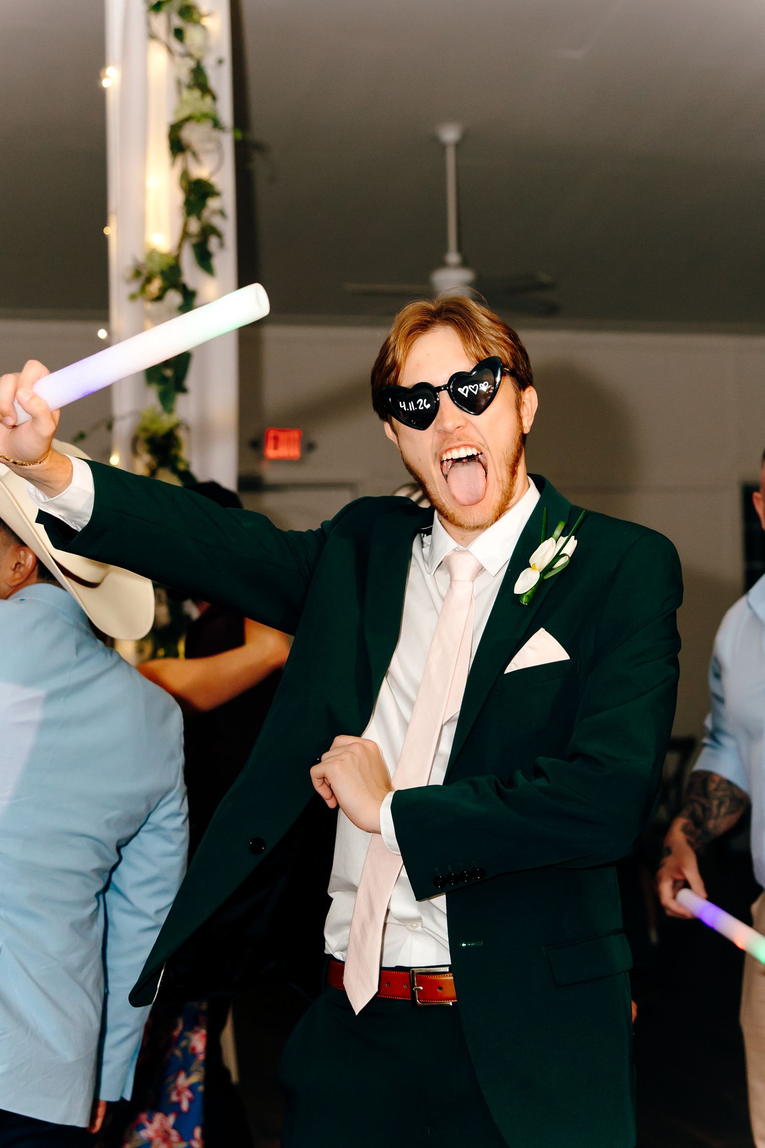 Groom in green suit and heart sunglasses dancing with glow stick at wedding reception.