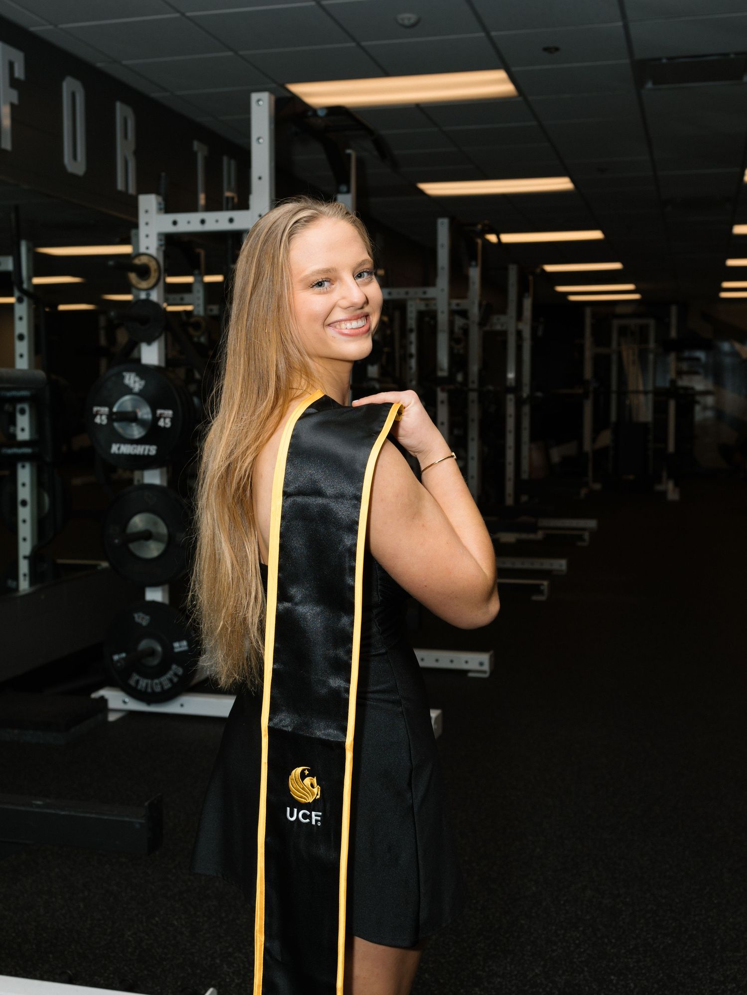 Smiling UCF graduate poses in black and gold graduation stole inside a gym facility.