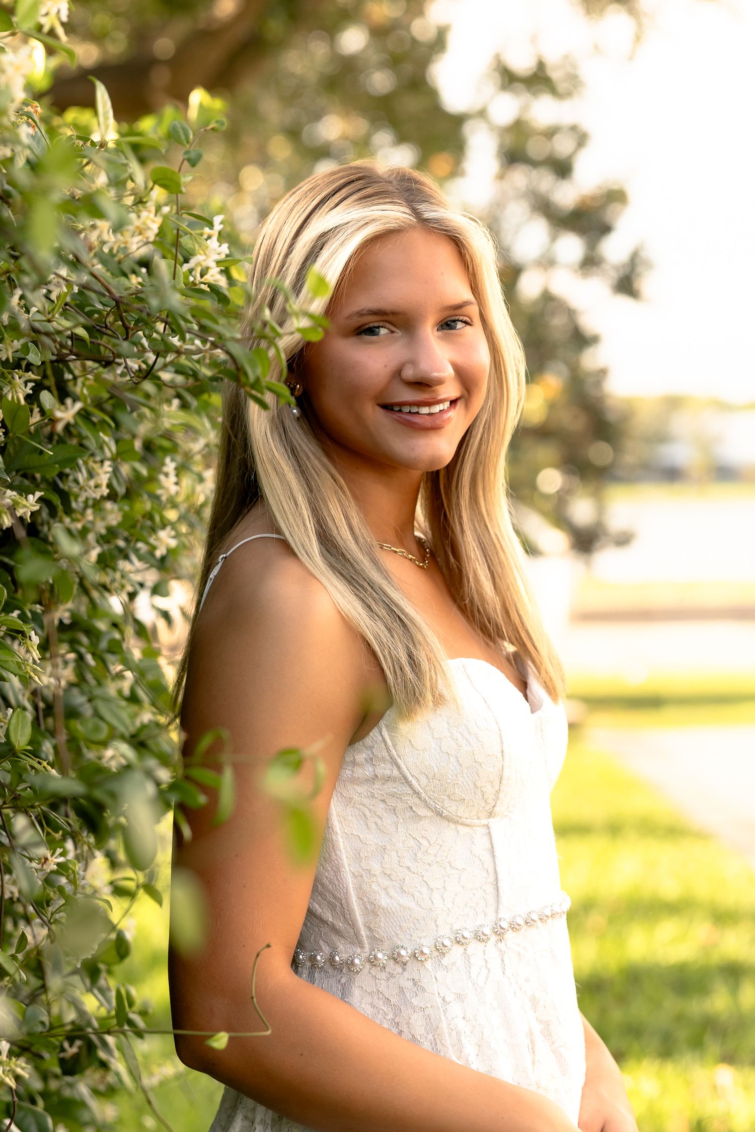 Smiling teenage girl in white lace dress standing by flowering bushes in golden hour sunlight.