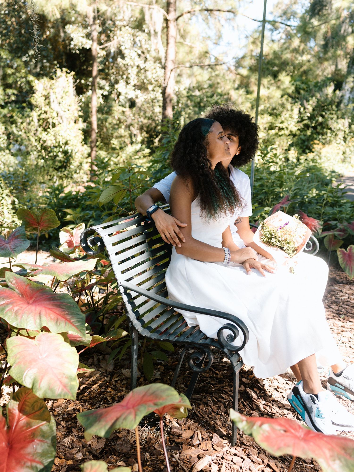 A romantic garden scene with someone in a white dress sitting on a vintage bench surrounded by lush greenery and pink flowers.