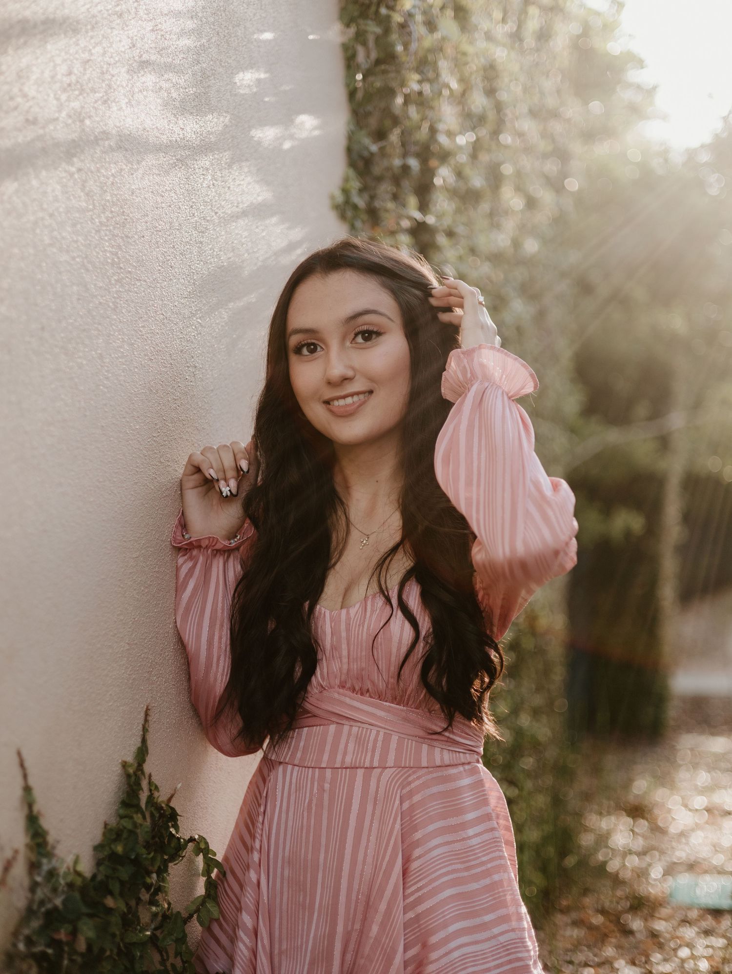 Portrait of someone in a pink pleated dress against a sunlit wall with soft shadows.