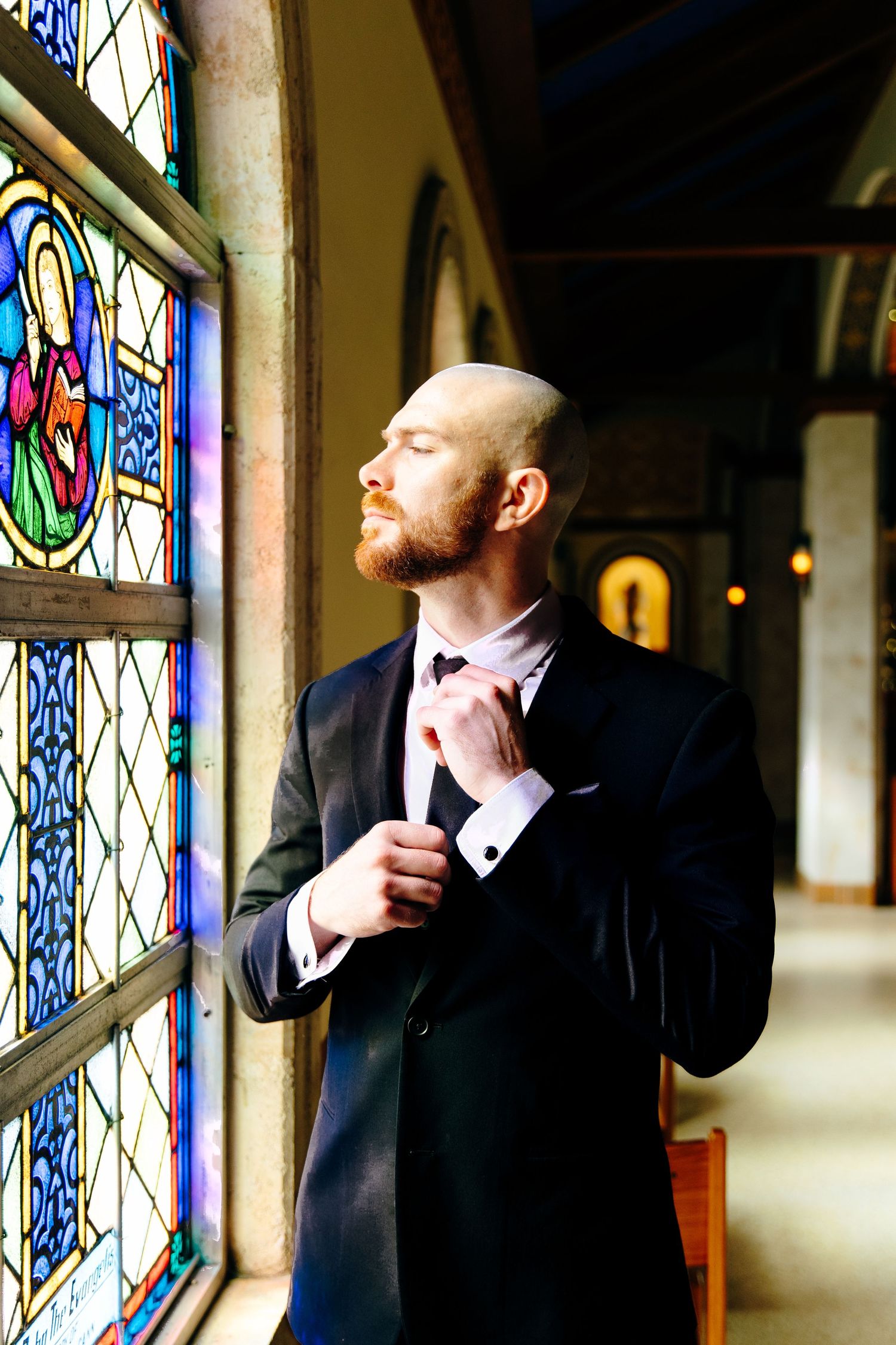 Dapper groom adjusts his bow tie beside colorful stained glass windows inside a historic church.
