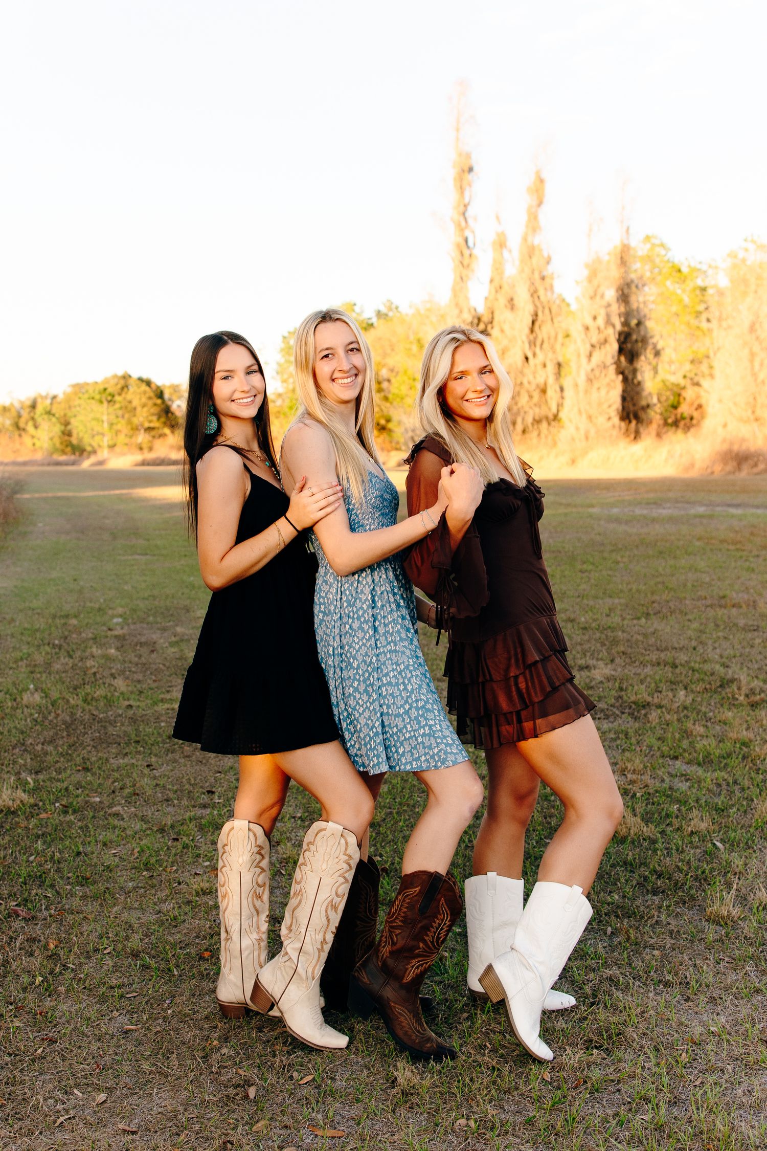 Three young women in mini dresses and cowboy boots pose together in a sunny field during golden hour.