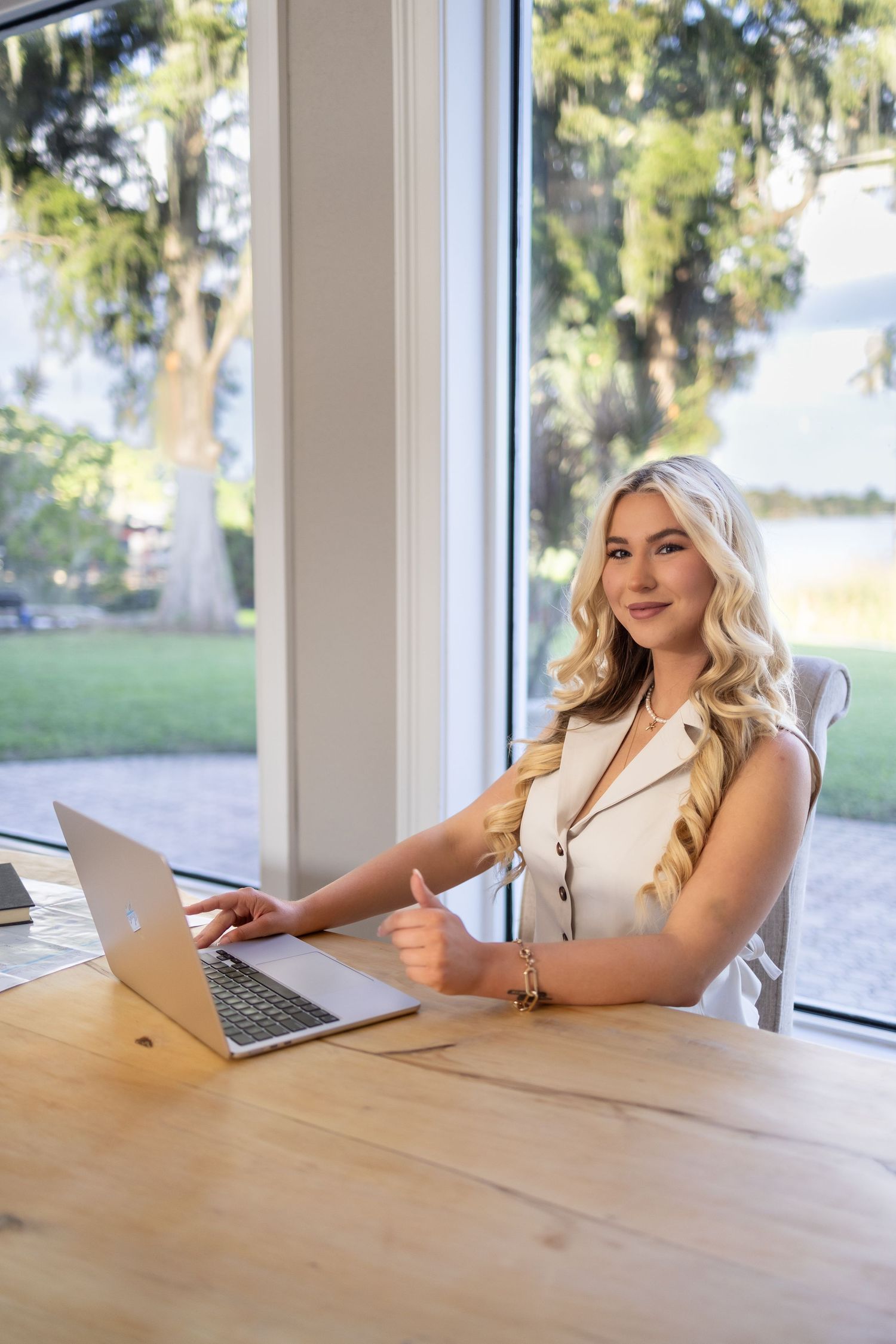 Young blonde woman working on laptop at wooden desk by large windows with garden view, giving thumbs up.
