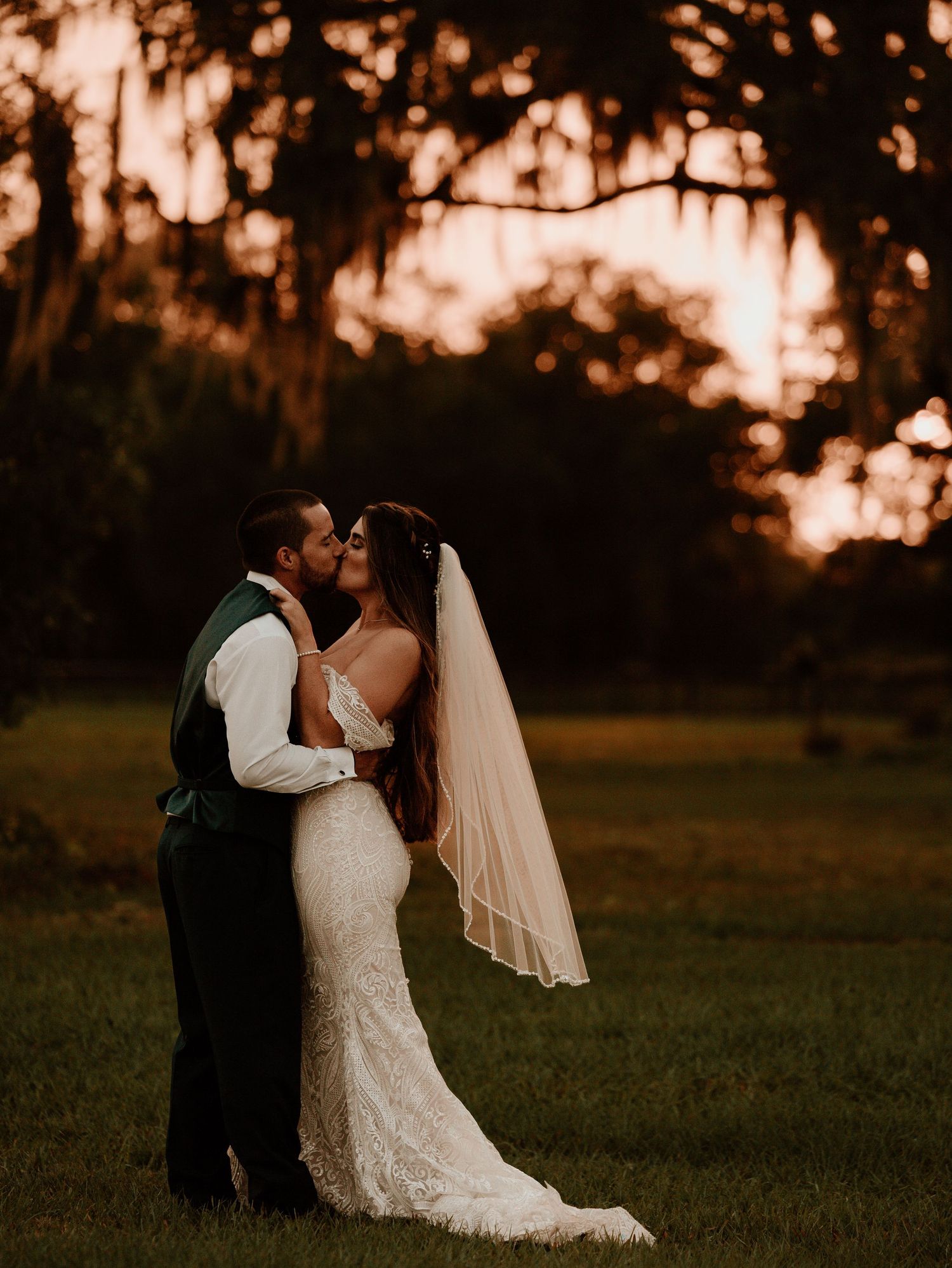 A couple shares a romantic moment at sunset in an outdoor wedding ceremony with golden light filtering through trees.