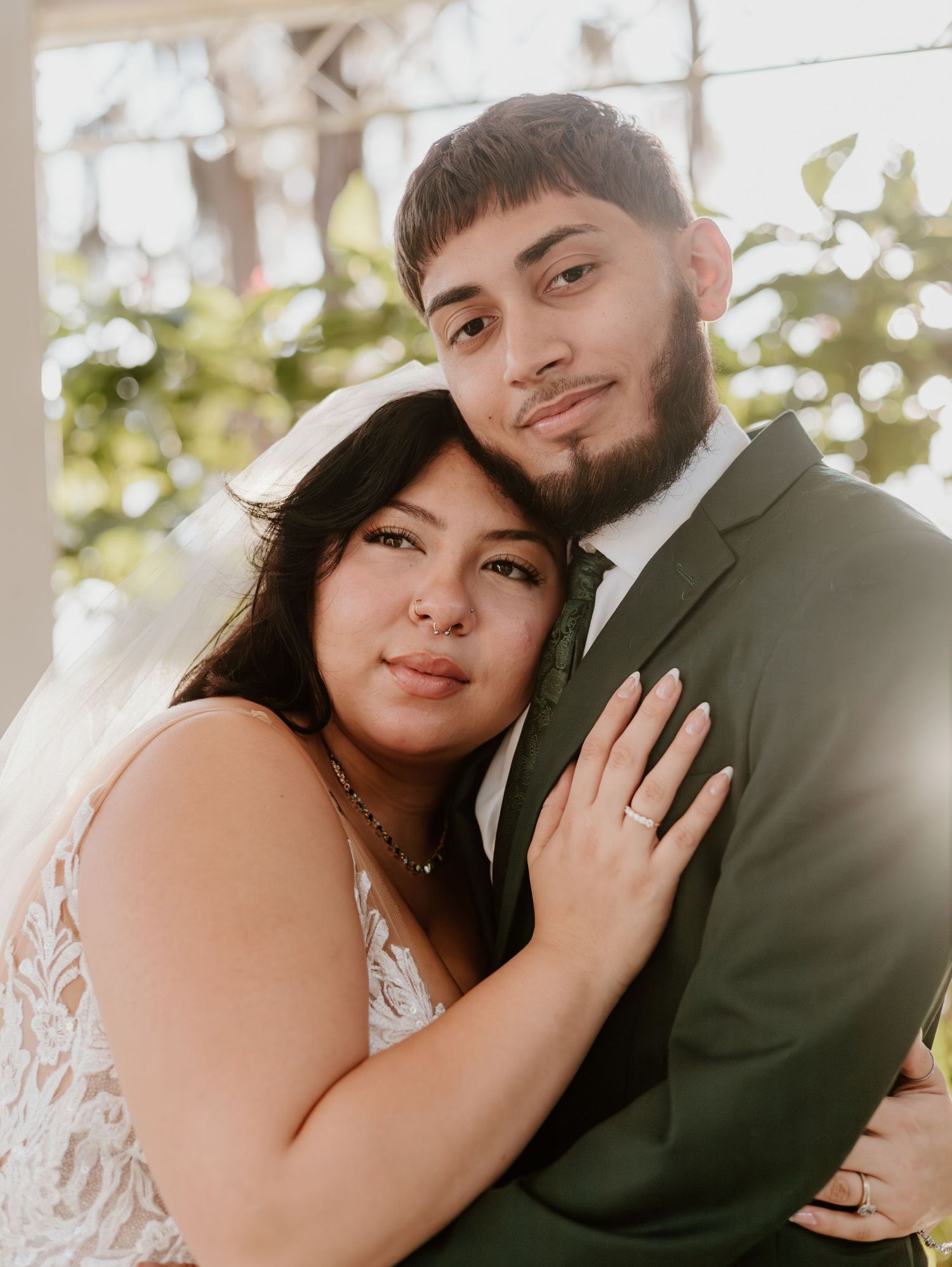 A couple embraces in wedding attire with the bride in a white lace dress and groom in an olive suit outdoors.