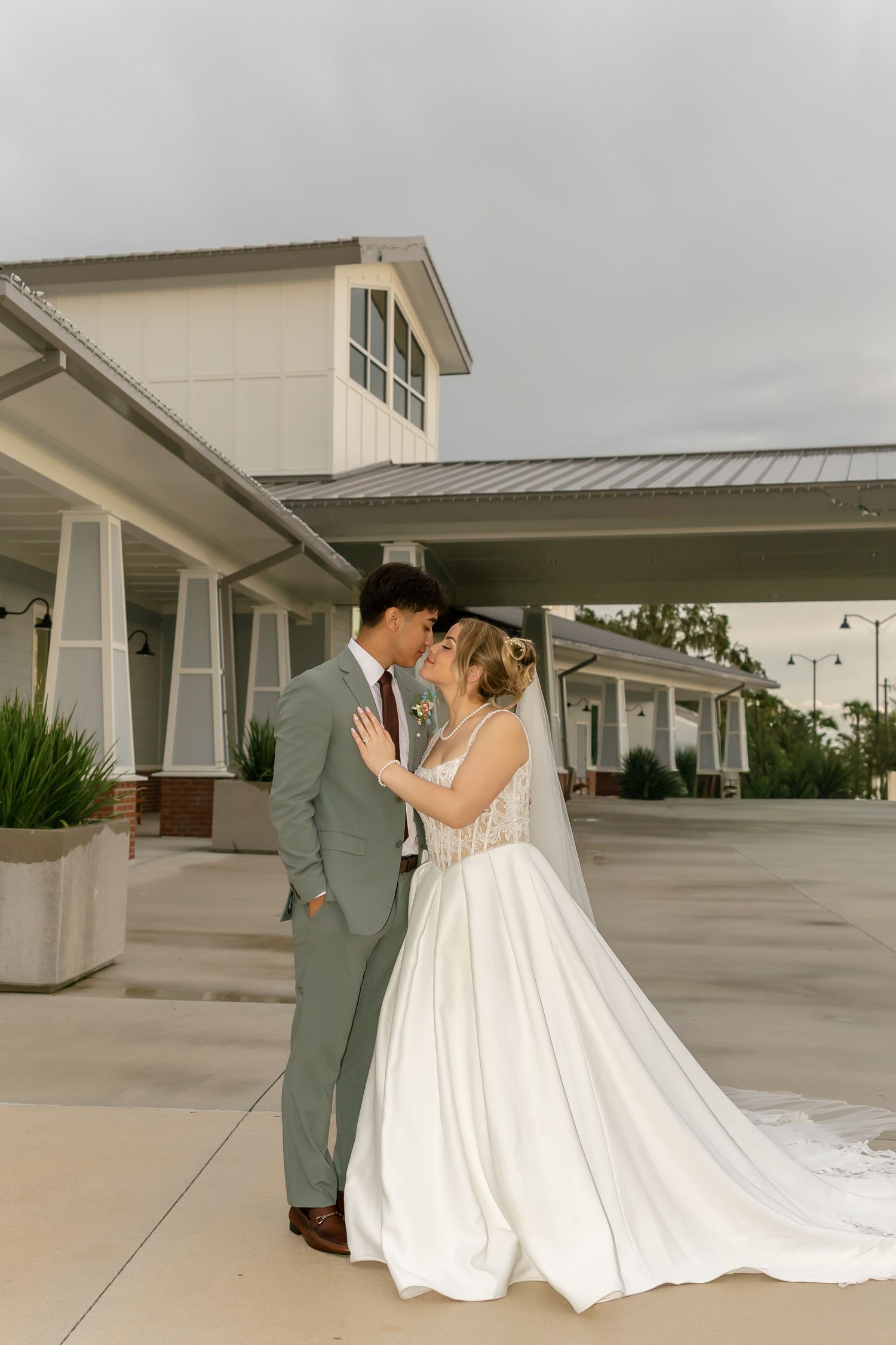 Bride and groom share a romantic kiss outside a white wedding venue, bride in flowing gown, groom in sage suit.