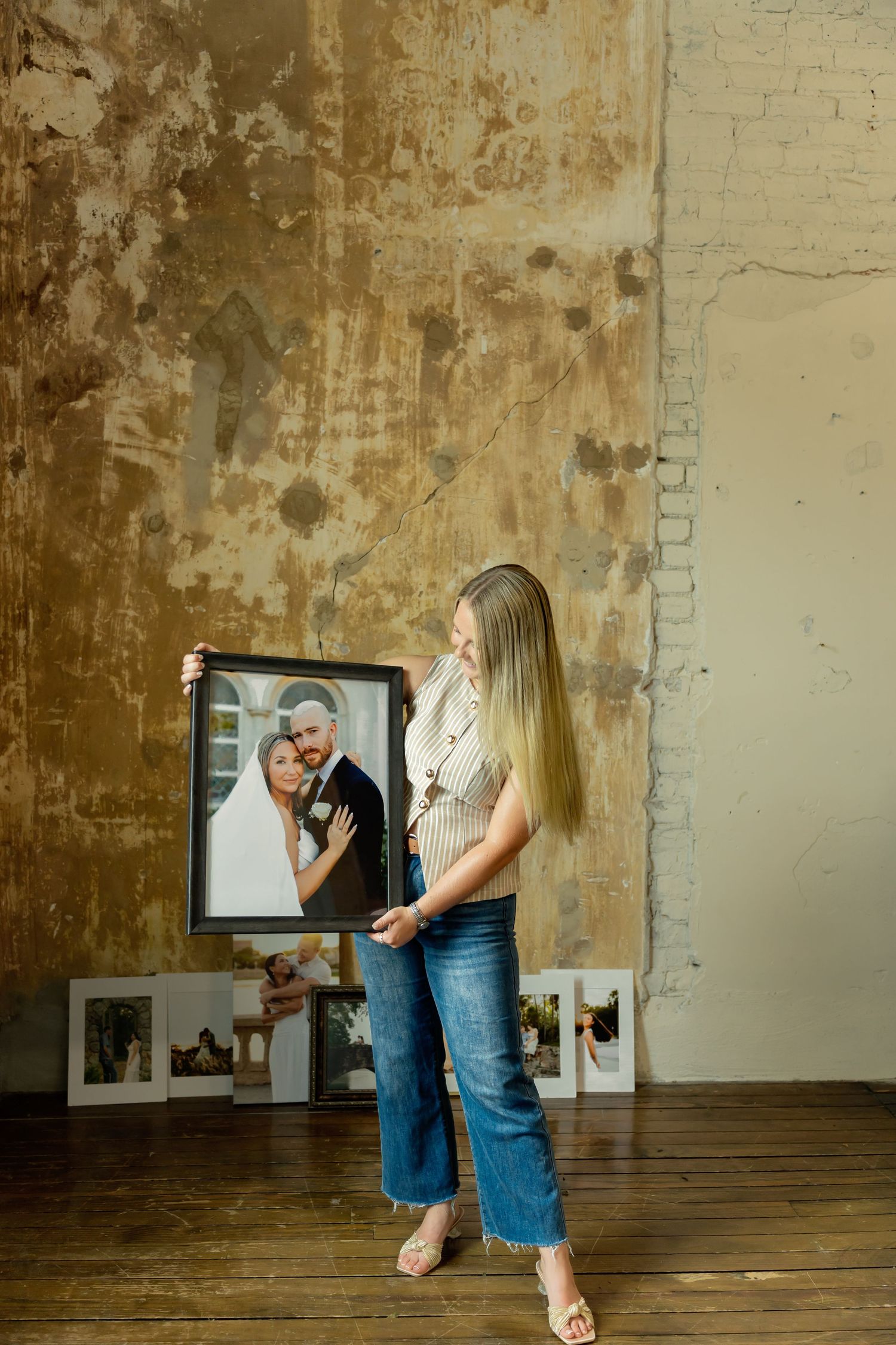 Woman holding a framed wedding portrait in a rustic studio with photos displayed on wooden floor.