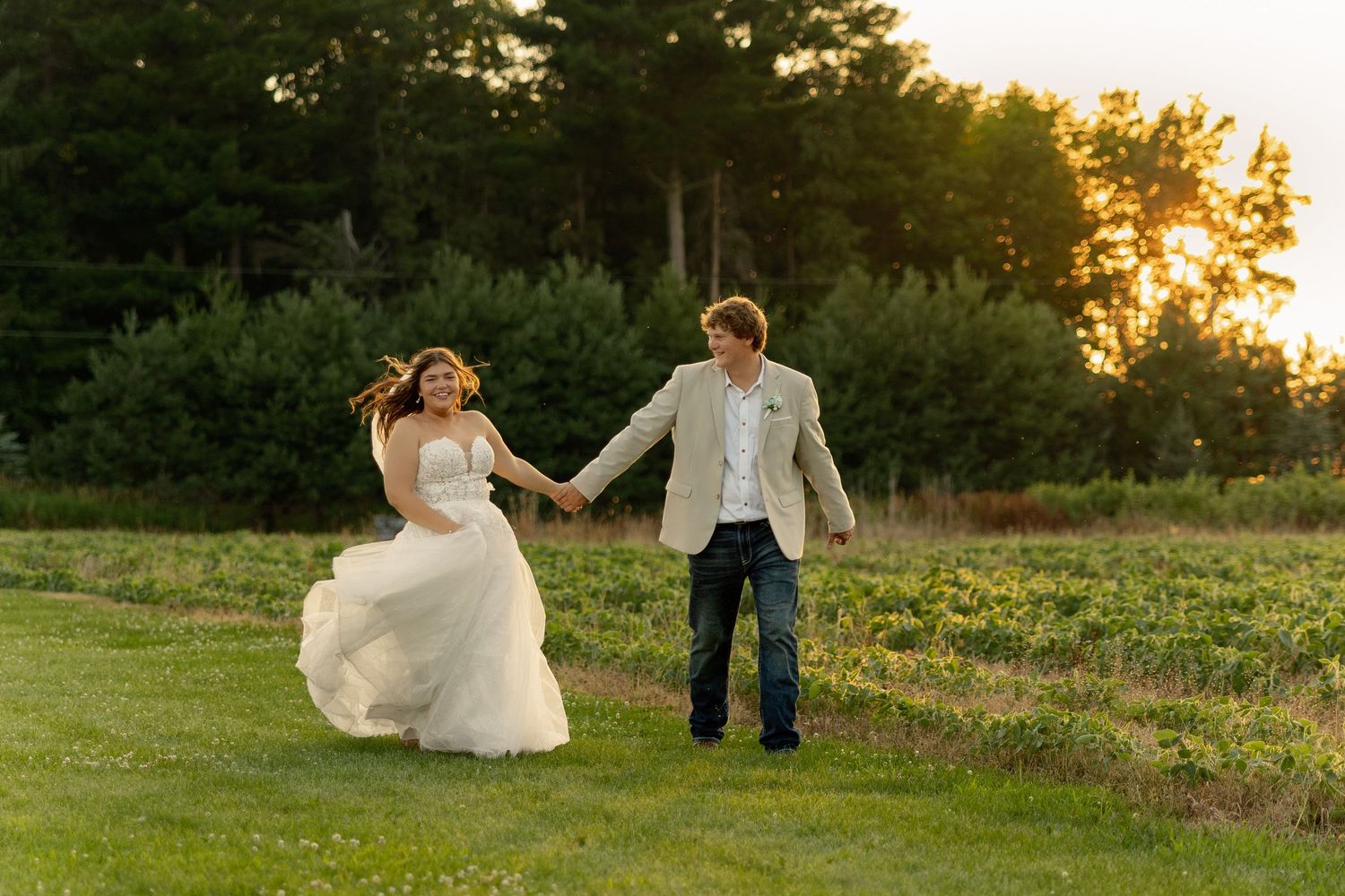 Bride in white gown and groom in jeans hold hands running through green field at golden sunset.