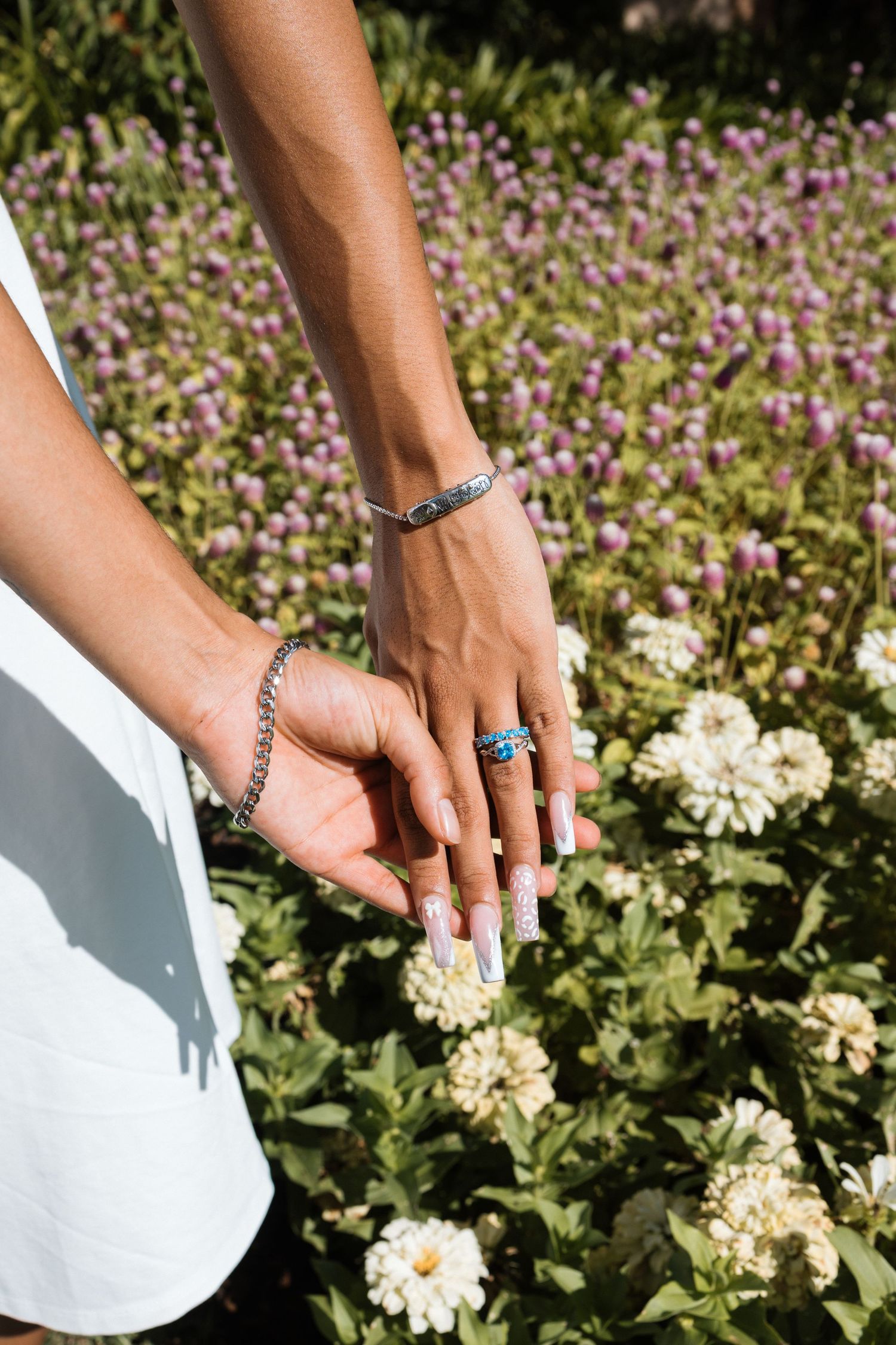 Elegant hands adorned with silver jewelry reach down to touch white flowers in a garden filled with purple blooms.