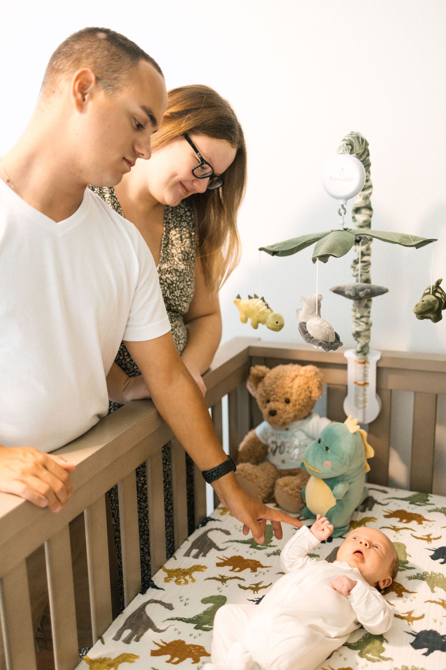 Young parents lovingly gazing at their newborn baby lying in a dinosaur-themed crib with stuffed animals and a mobile.