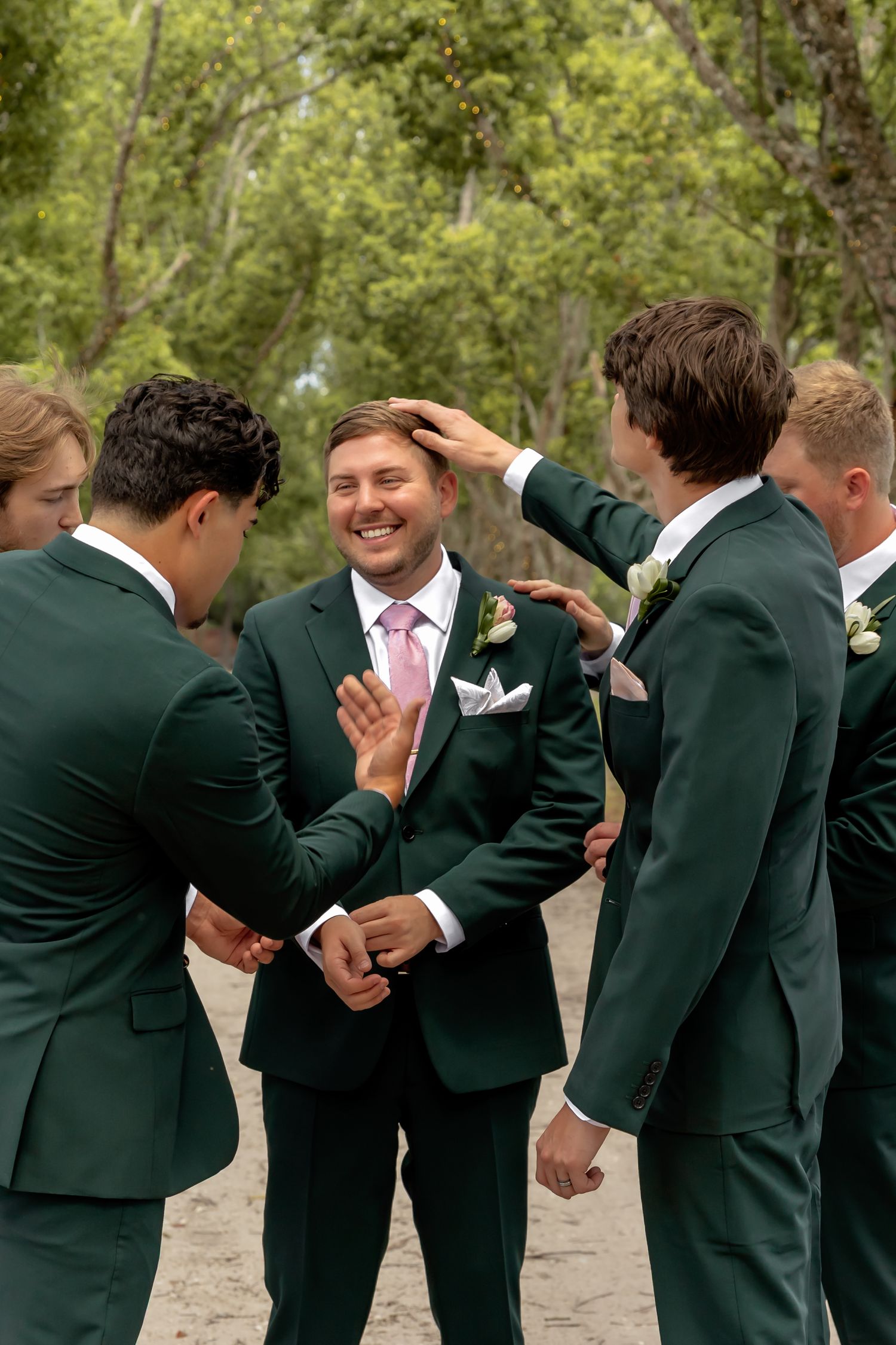 Groom in dark green suit smiles as groomsmen adjust his boutonniere and hair outdoors.