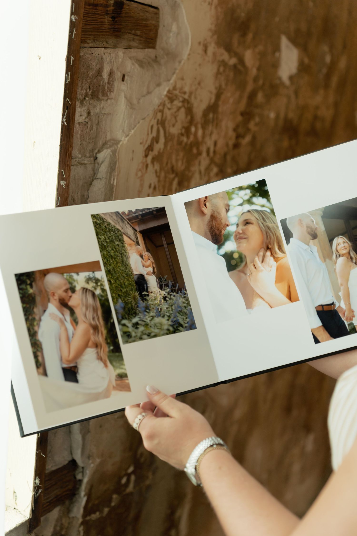 Hands holding an open wedding photo album displaying romantic couple portraits against a rustic stone wall backdrop.