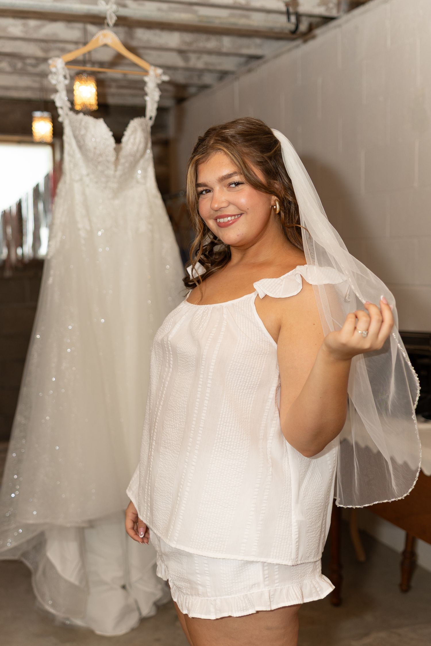 Bride in white shorts set holding veil, with sparkly wedding gown hanging in background.