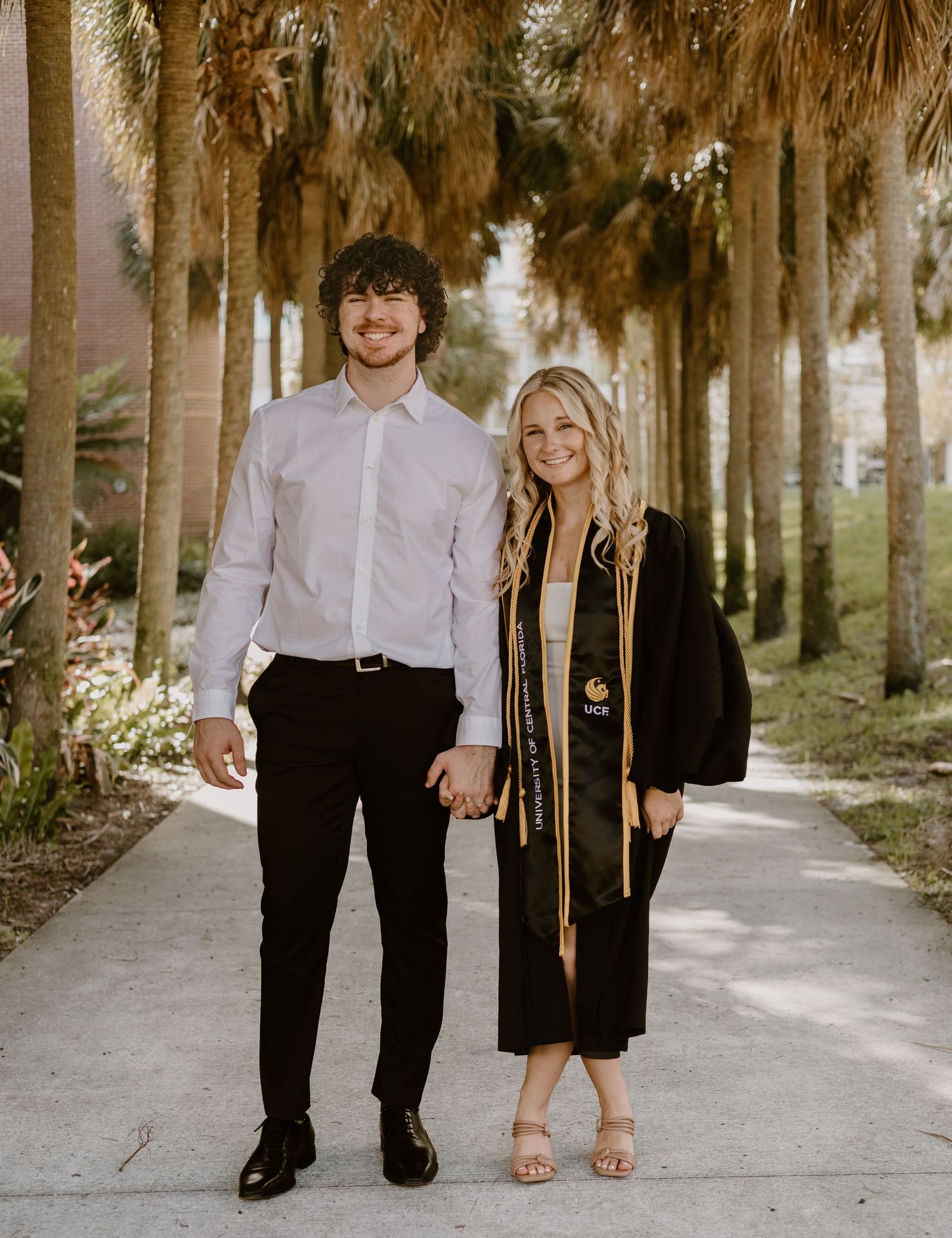 A smiling couple poses on a palm-lined walkway, the woman wearing a black graduation gown with gold stole and cords.
