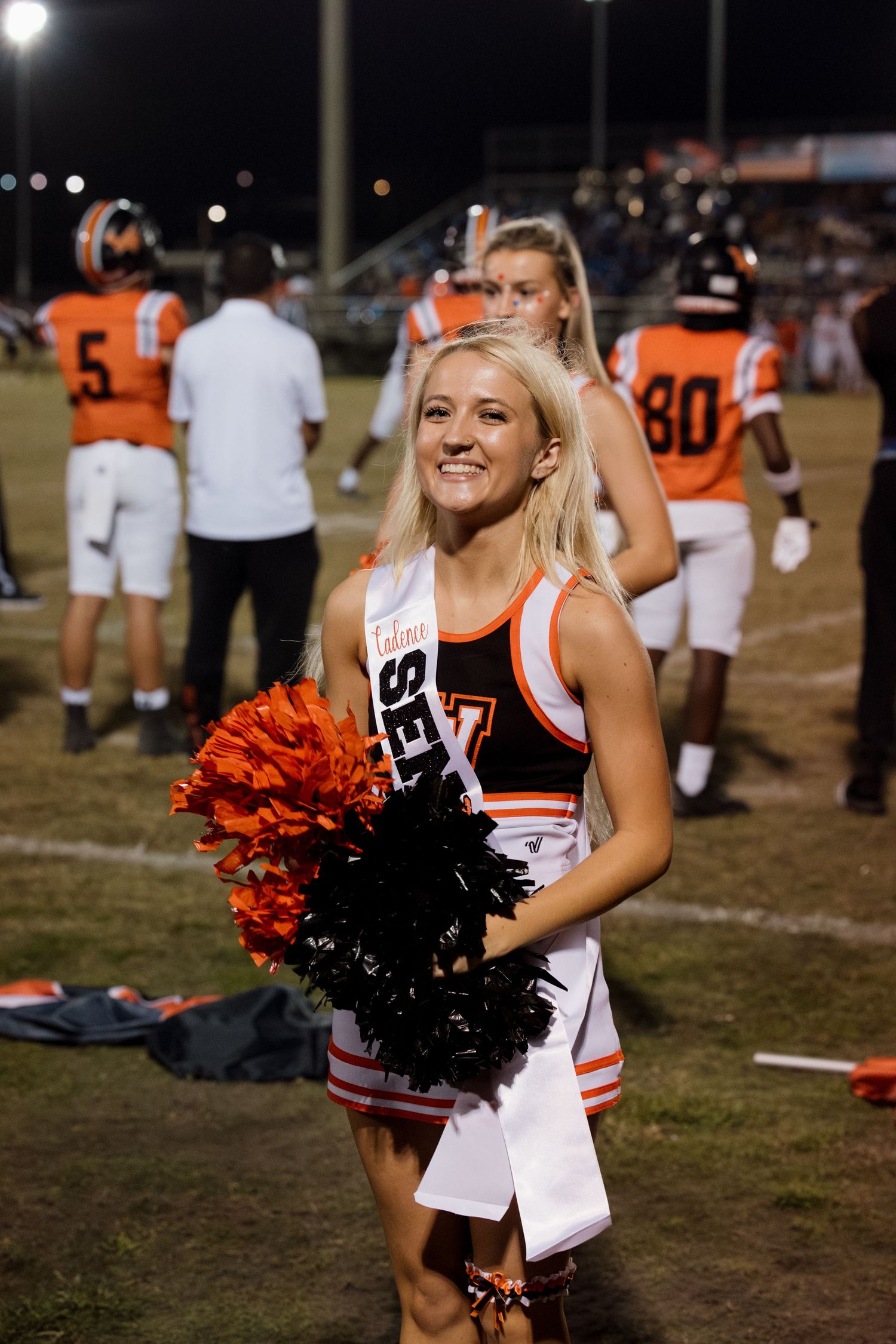 Cheerleaders in red and white uniforms with black pom poms stand at the sidelines of a nighttime football field.